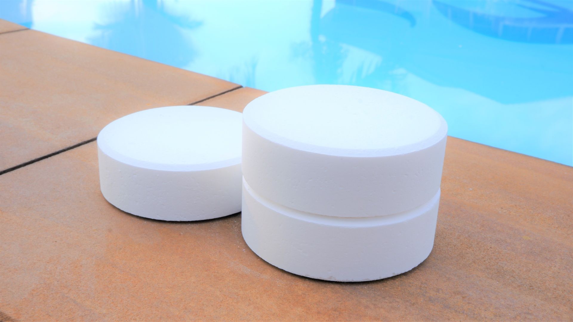 Two white chlorine tablets rest on a poolside deck beside clear blue water.