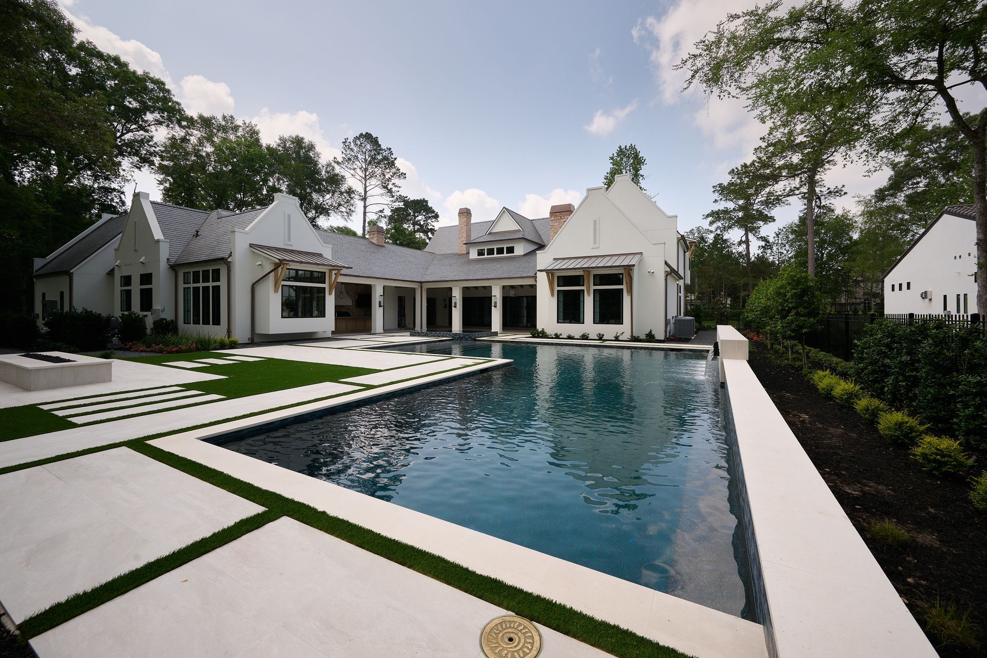 A white house with a metal roof overlooks a rectangular swimming pool surrounded by a geometric stone and grass patio.
