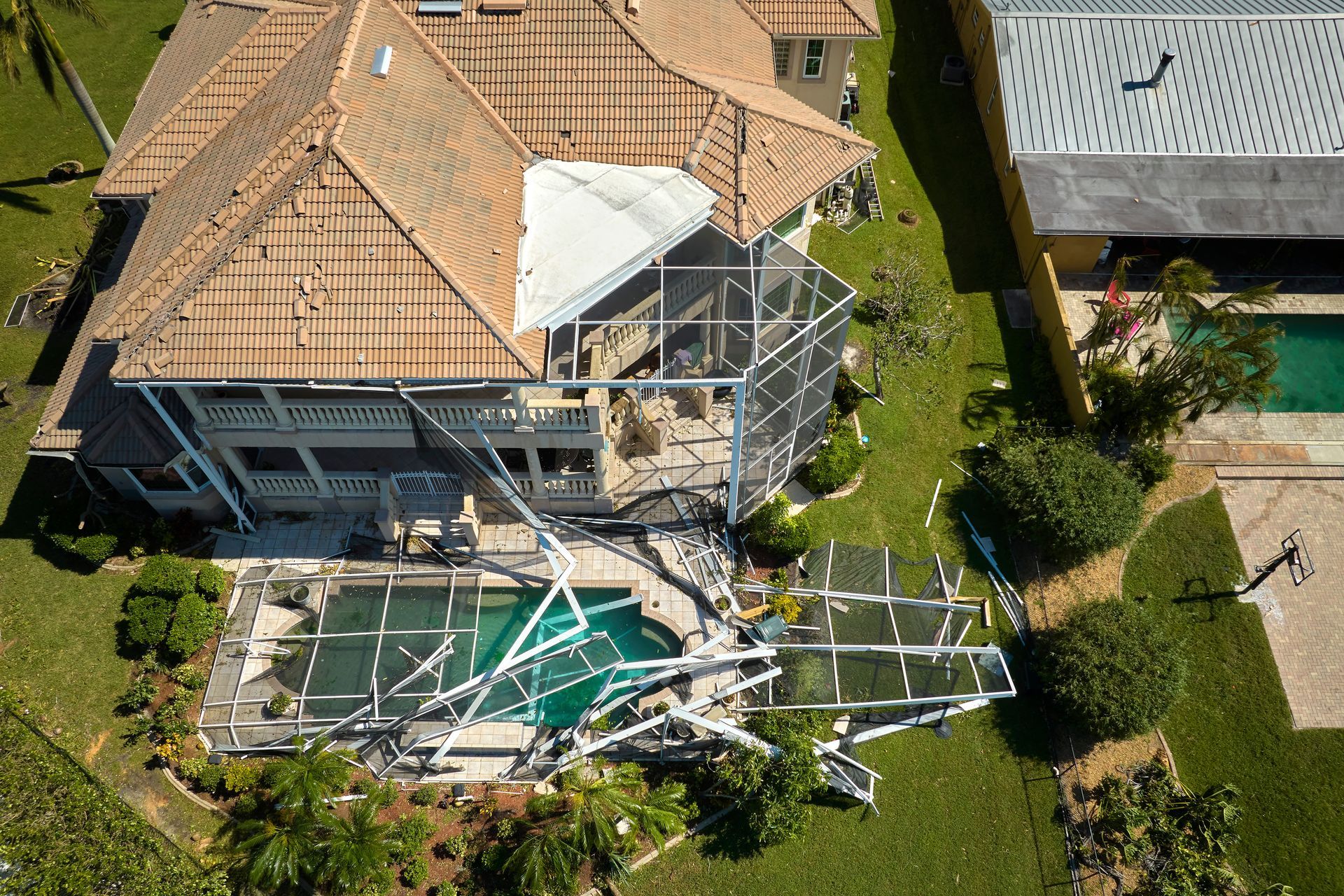 An aerial view of a house with a damaged roof and a collapsed metal pool enclosure in the backyard.