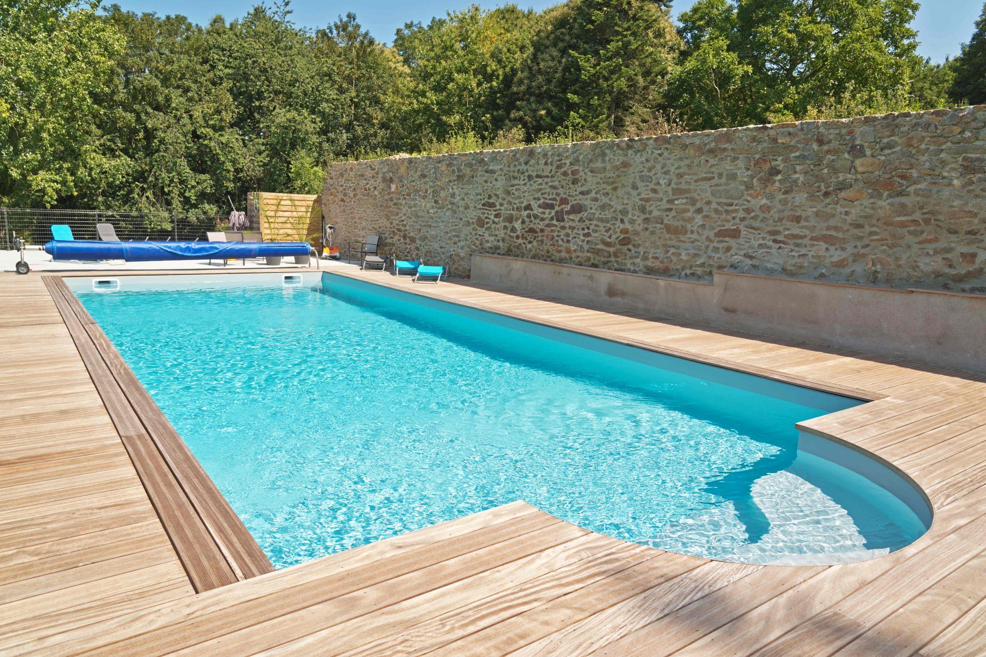 Outdoor swimming pool with blue water, wooden deck, and stone wall in the sun