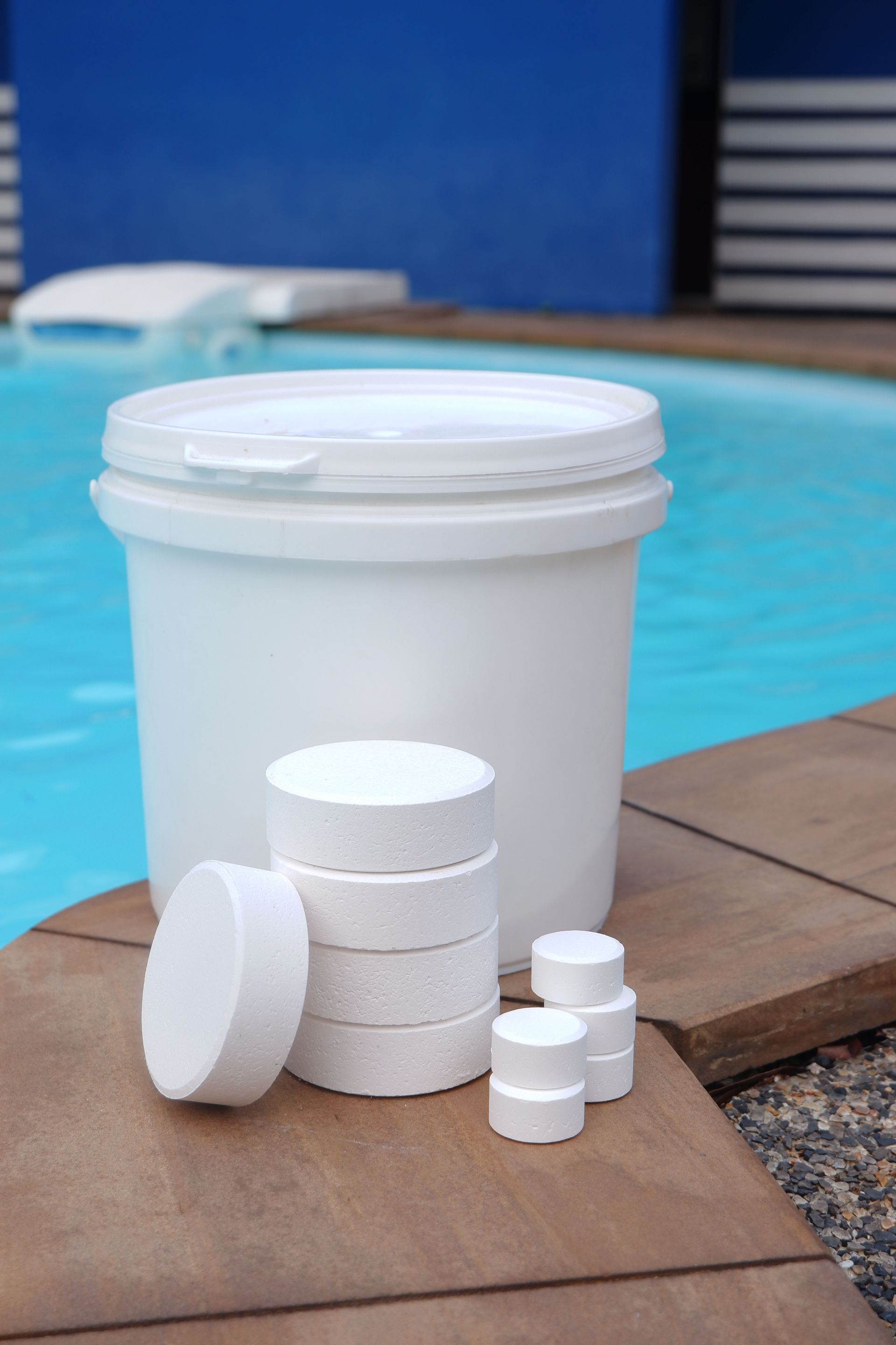 A white plastic bucket and stacks of pool chlorine tablets on a tiled pool deck beside clear blue water.