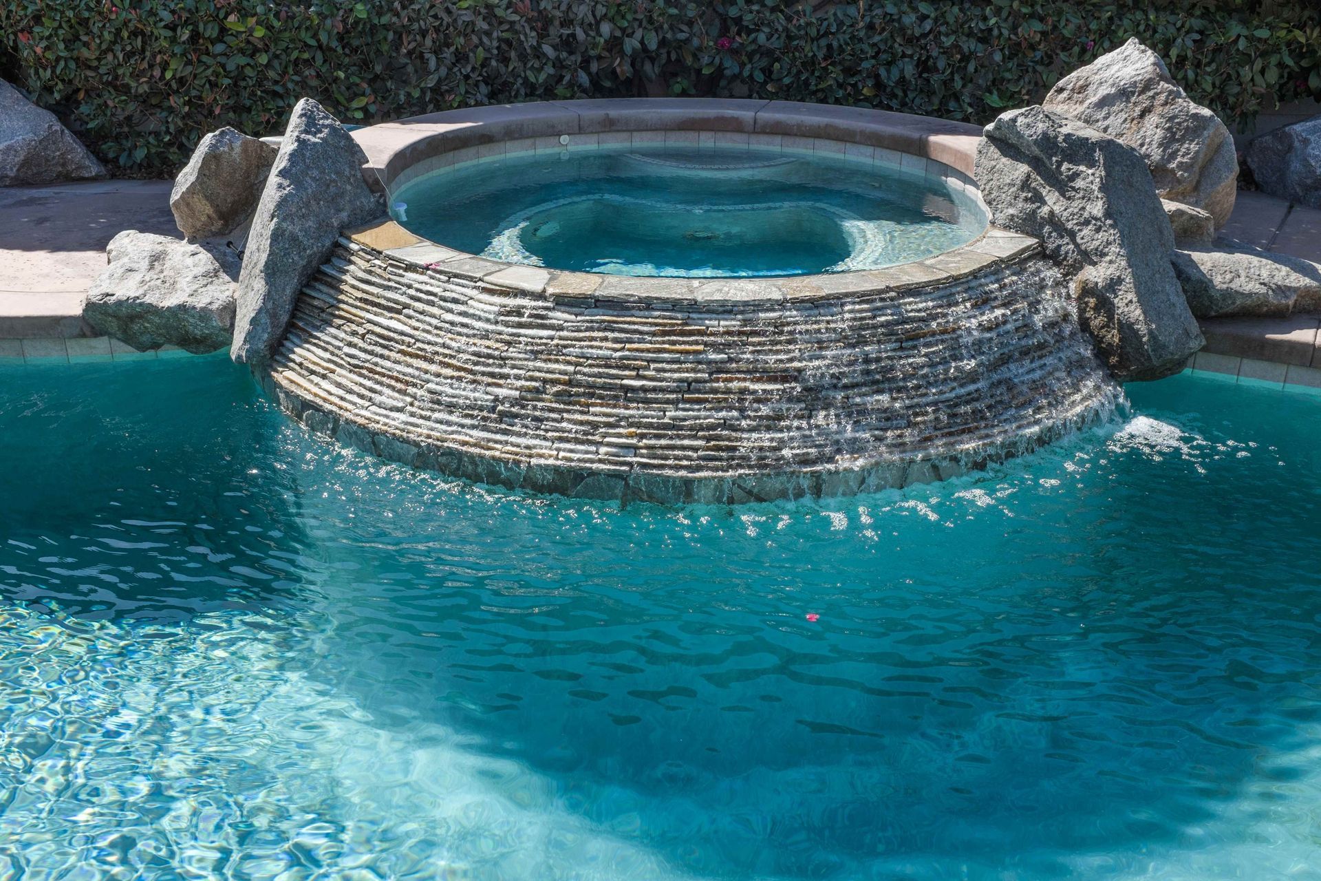 Circular stone hot tub beside a turquoise swimming pool with rocks around it.