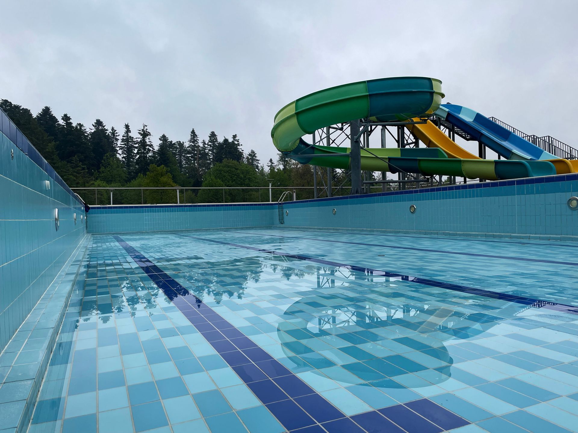 An empty outdoor swimming pool with lane markers, featuring a large, multi-colored winding water slide in the background.