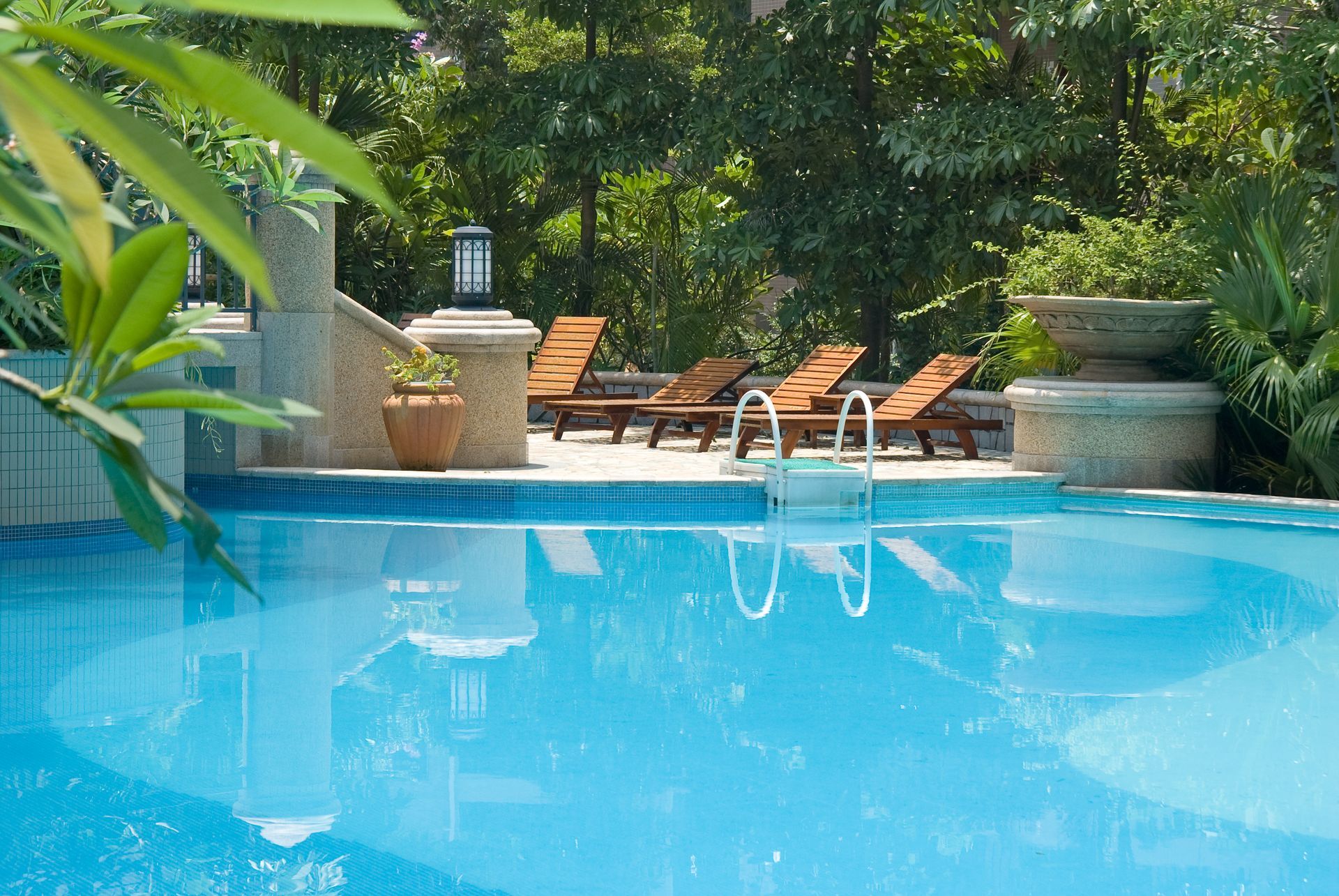 A sunny swimming pool surrounded by lush greenery, featuring wooden lounge chairs on a stone deck.