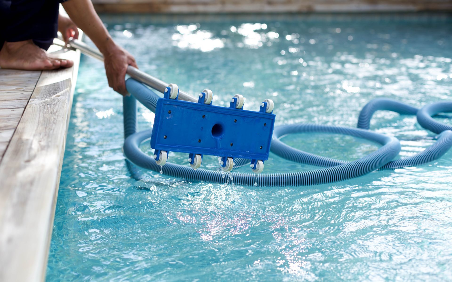 A person uses a blue pool vacuum head attached to a pole to clean a swimming pool.