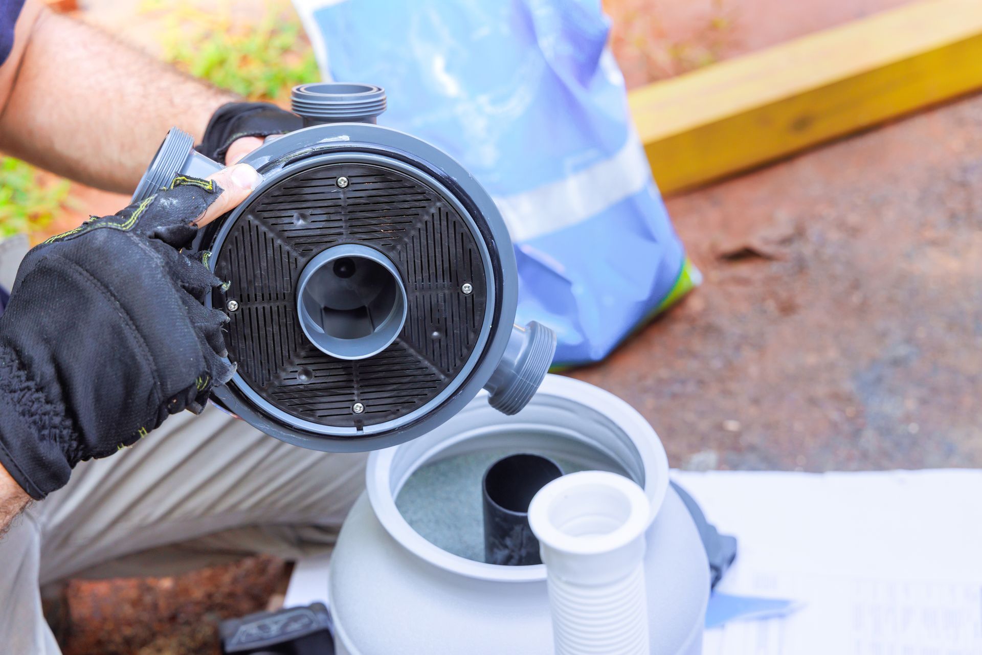 A person wearing gloves holds a circular black pool pump filter component above an open, light-colored filter canister.