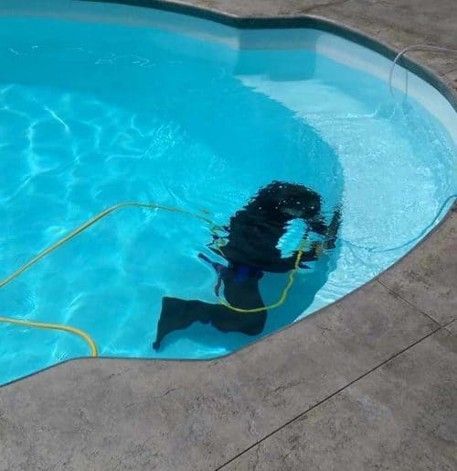A person in dark clothing and a snorkel mask is underwater in a light blue swimming pool, holding a yellow hose.