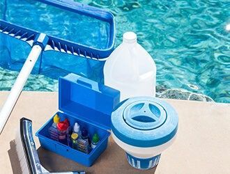 Pool cleaning tools, including a net, a test kit box, a chemical dispenser, and a jug, sit on a deck by a blue pool.