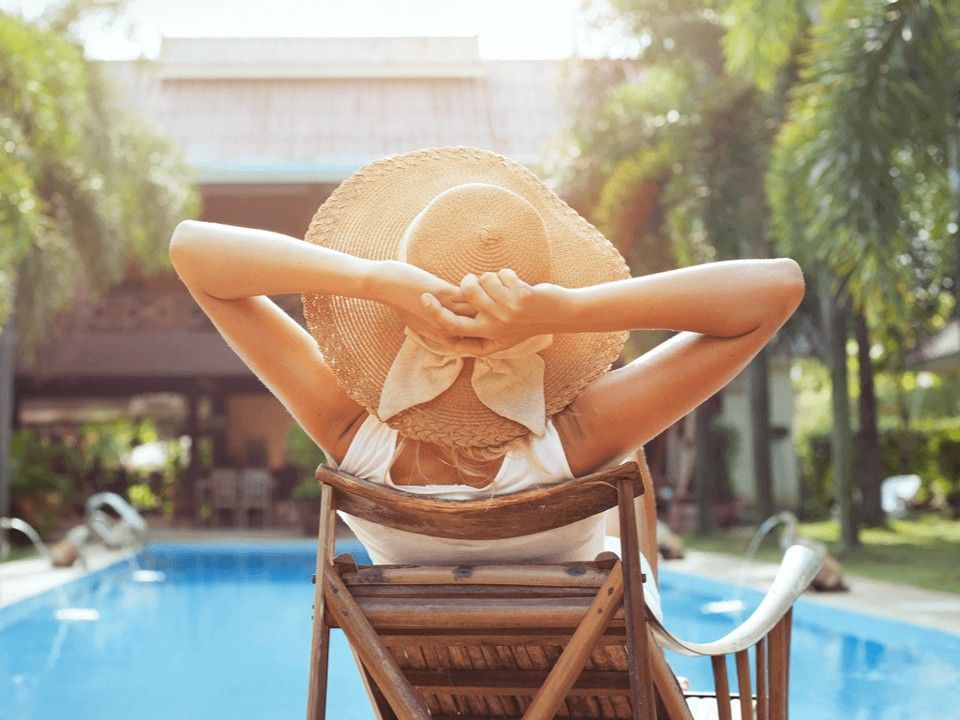 A person wearing a wide-brimmed straw hat relaxes in a wooden chair overlooking a sunlit swimming pool.
