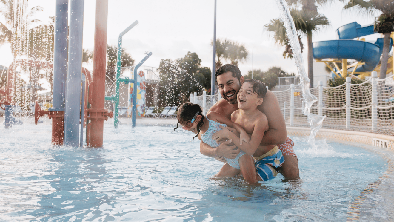 A smiling adult hugs two children in a splash park as water sprays around them.