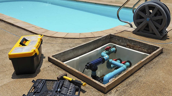 A yellow toolbox and open tool kit sit next to an open inground pool pump housing on a concrete pool deck.