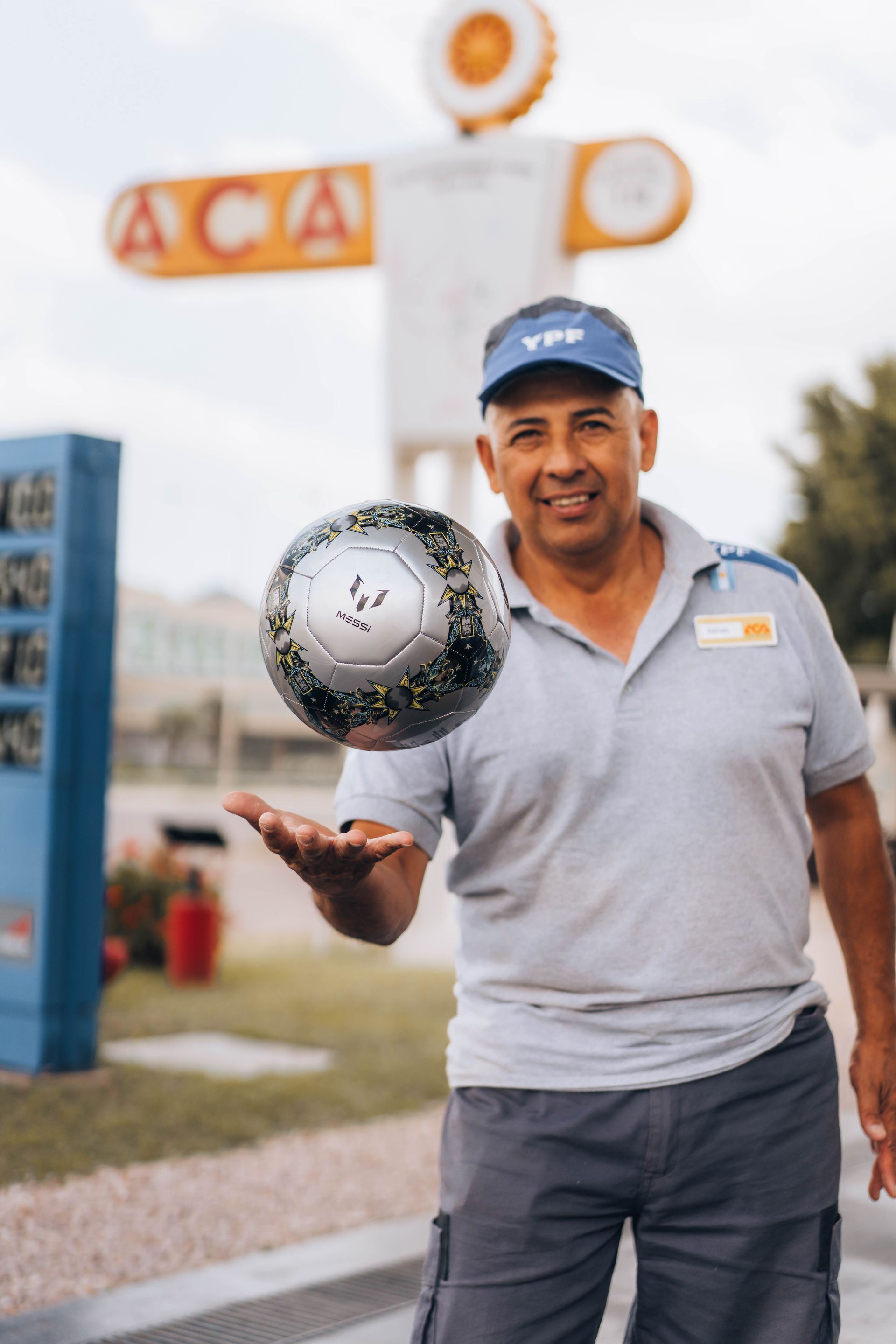Man in uniform holding a soccer ball outside a gas station. He smiles, wearing a blue hat.