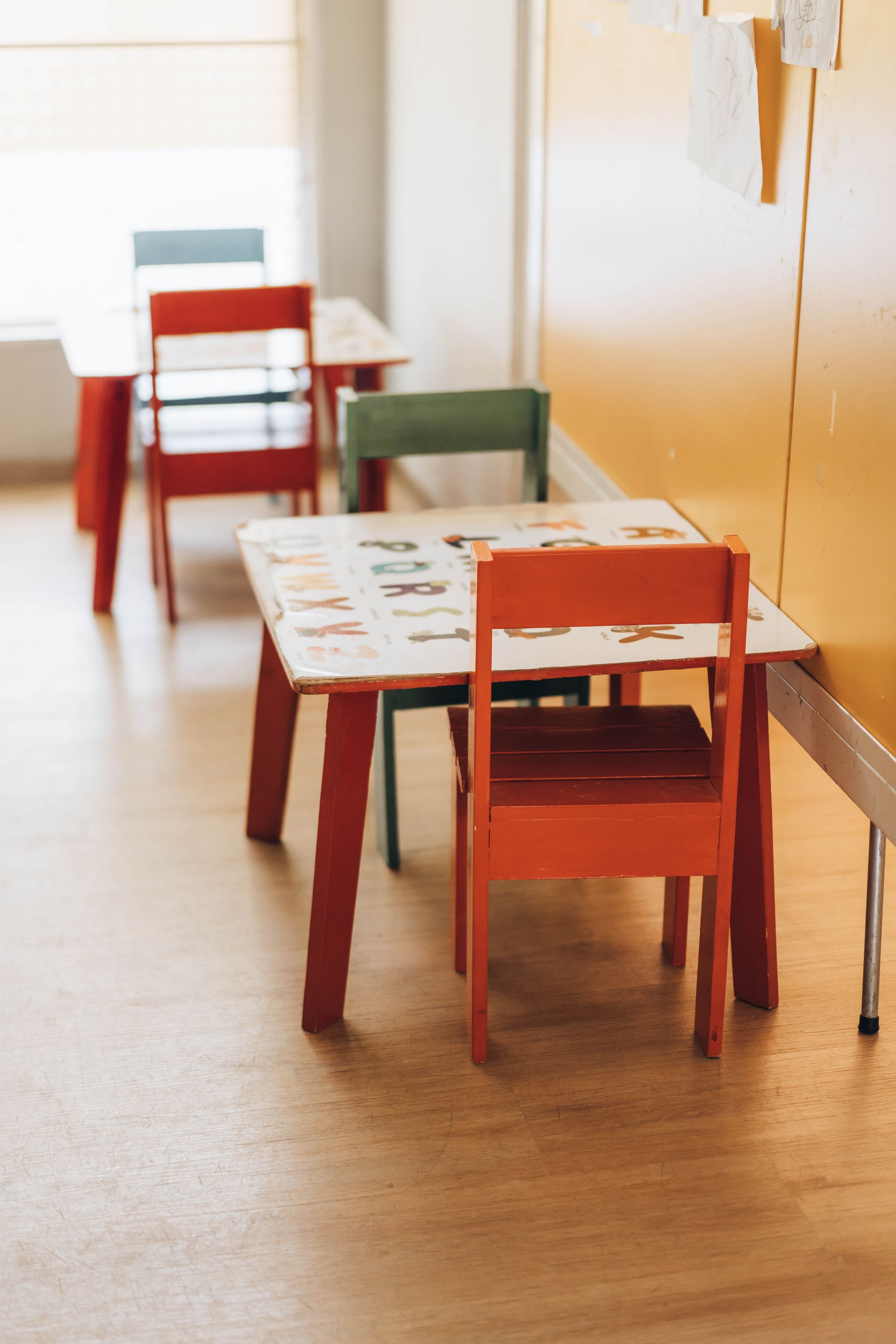 Red and green children's tables and chairs lined up in a bright room, near a window and yellow wall.