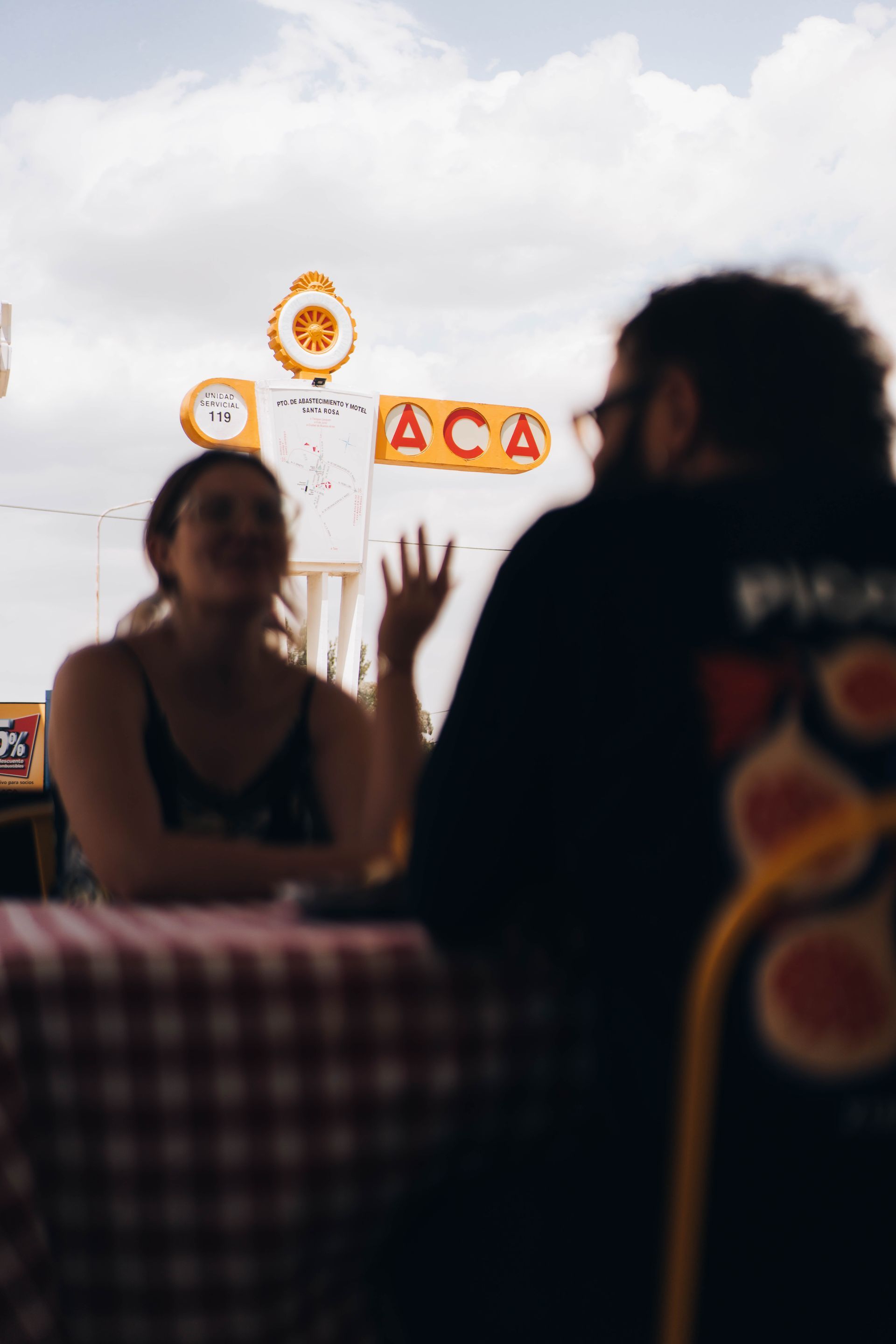 Two people at a table with a red checkered tablecloth, under a sign that reads