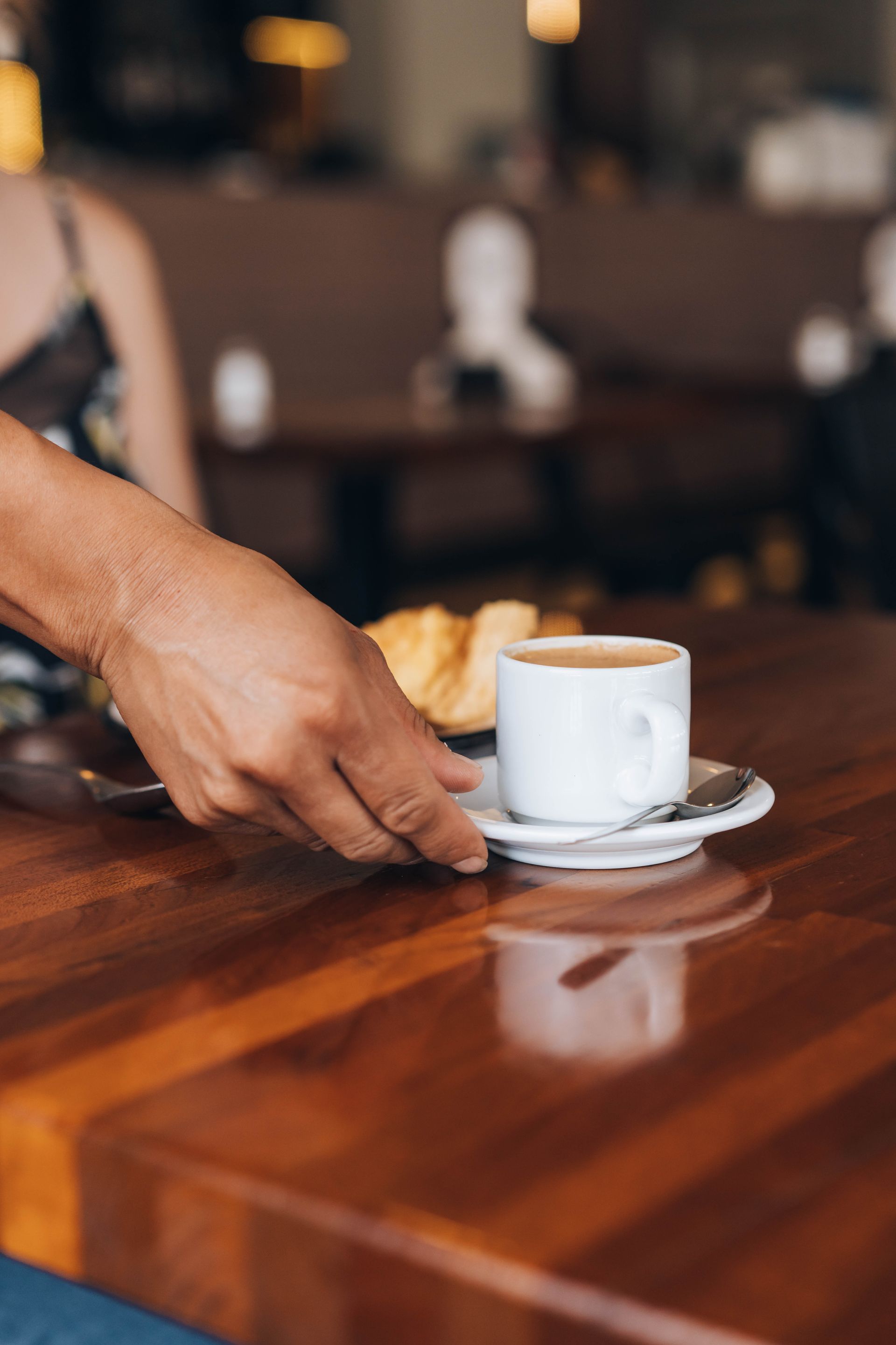 Hand sets down a coffee cup with saucer on a wooden table in a cafe.