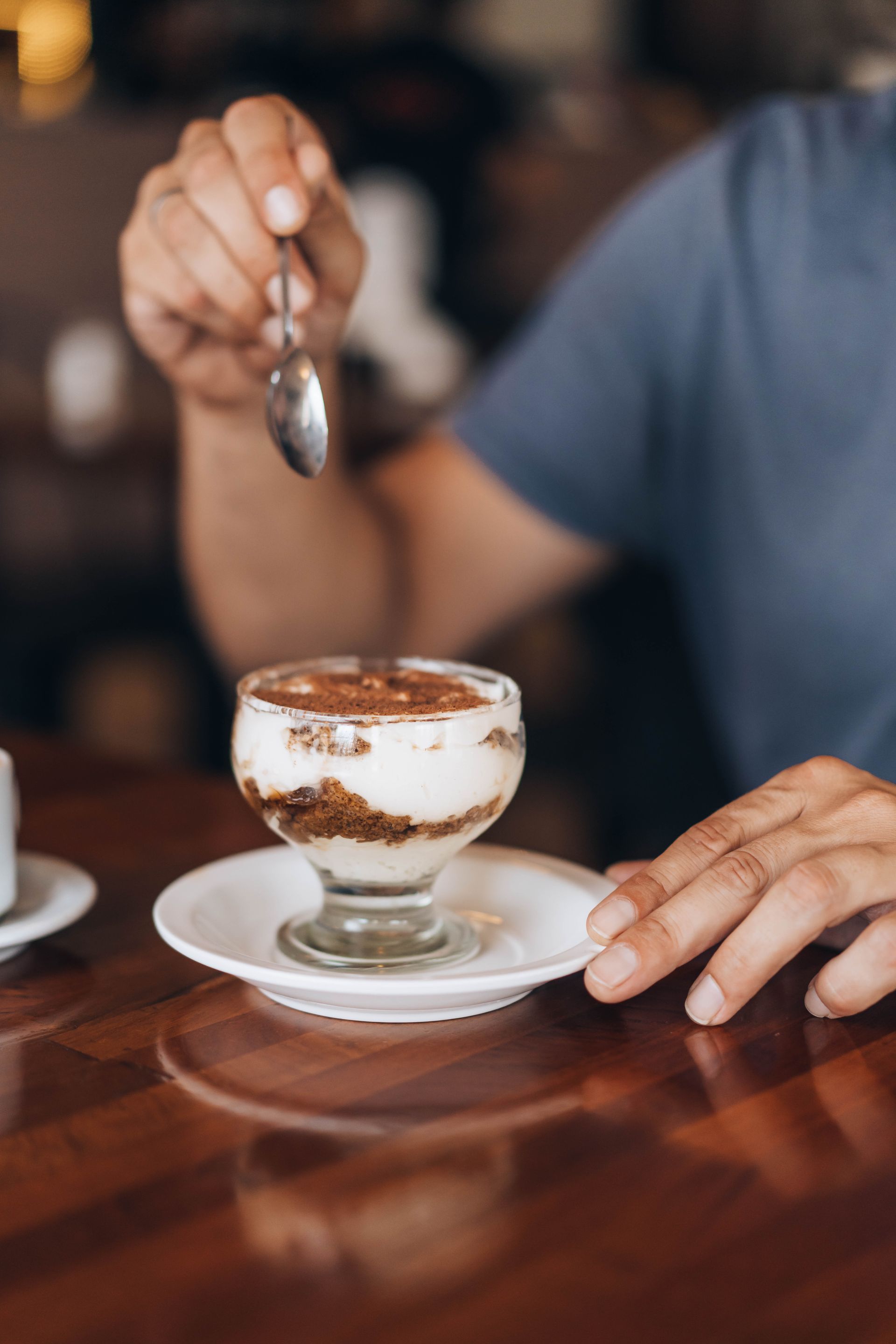 Person enjoying tiramisu in a glass, holding a spoon, on a wooden table.