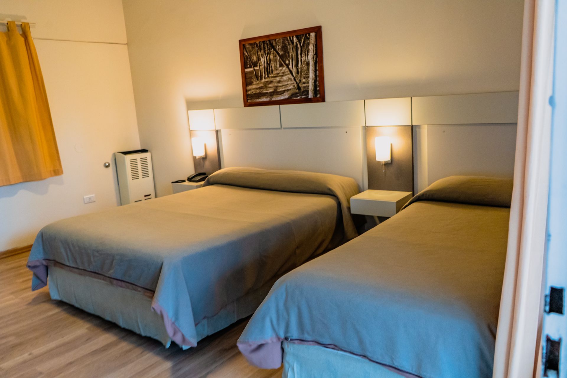 A hotel room with two beds; a double and a single, wall lights, and a landscape painting.