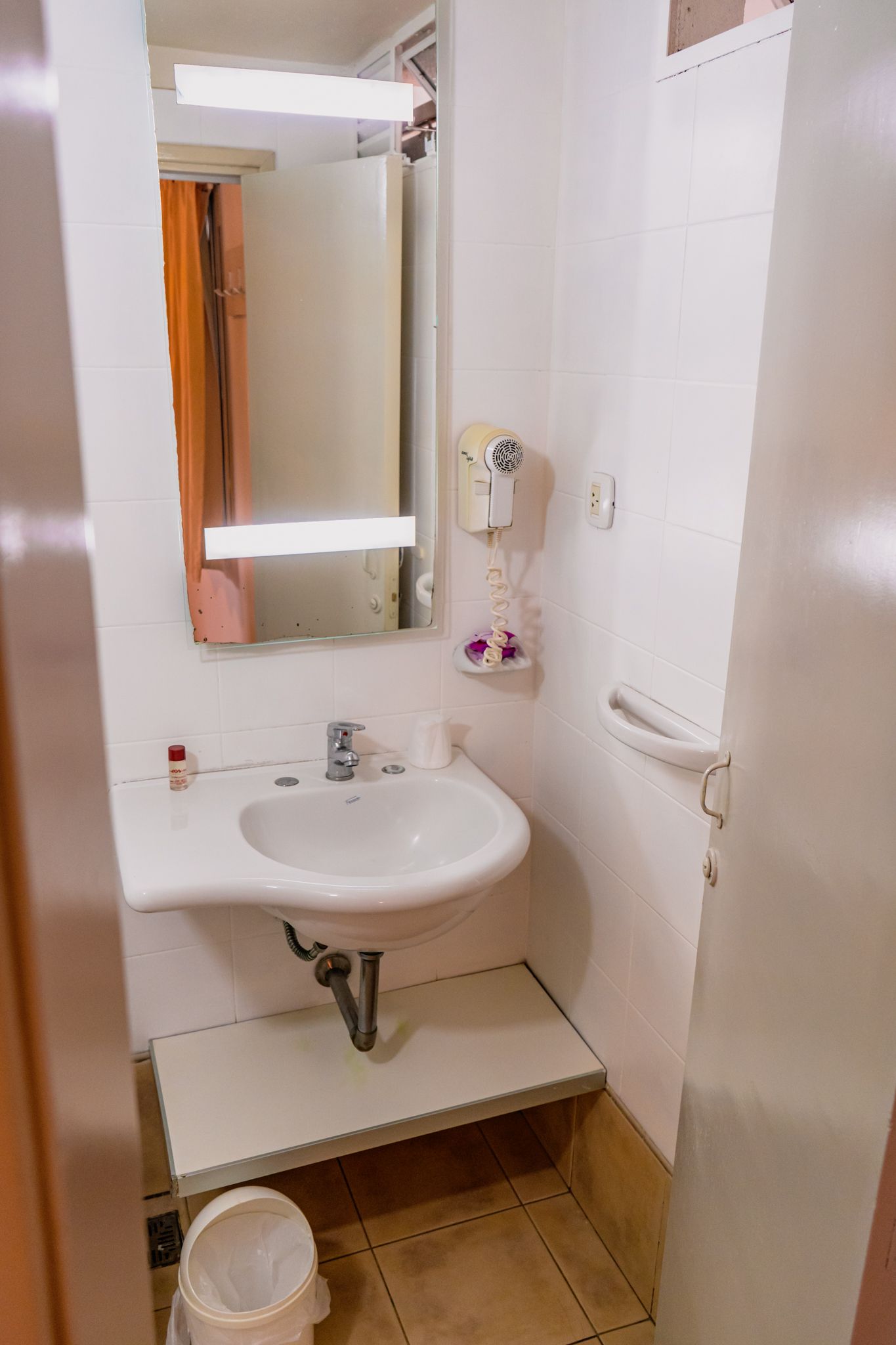 Small bathroom with a sink, mirror, hair dryer, and toilet. White walls and a tan floor.