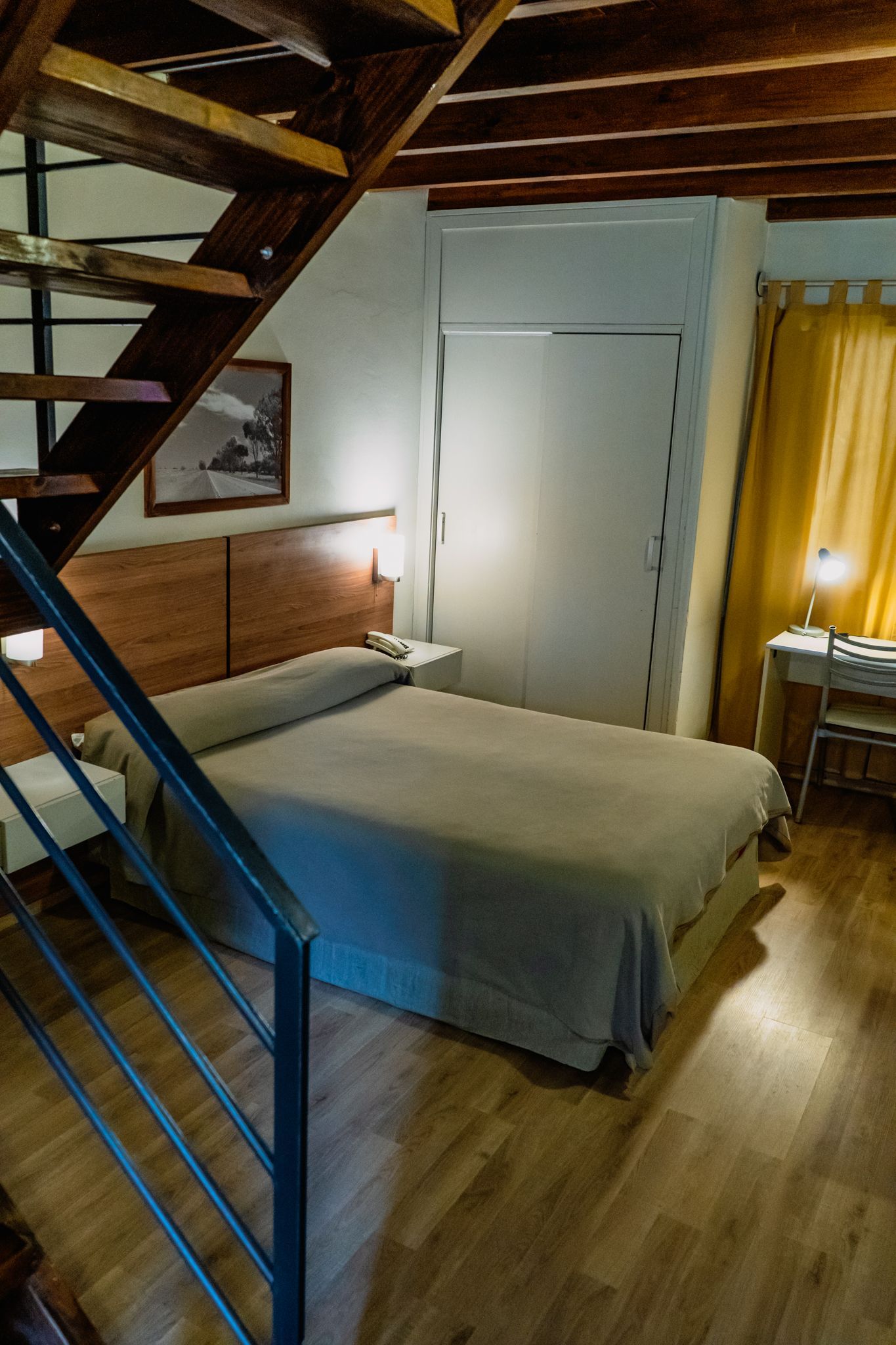 Bedroom with wooden staircase, bed, and desk. Yellow curtains, brown wood, neutral colors.