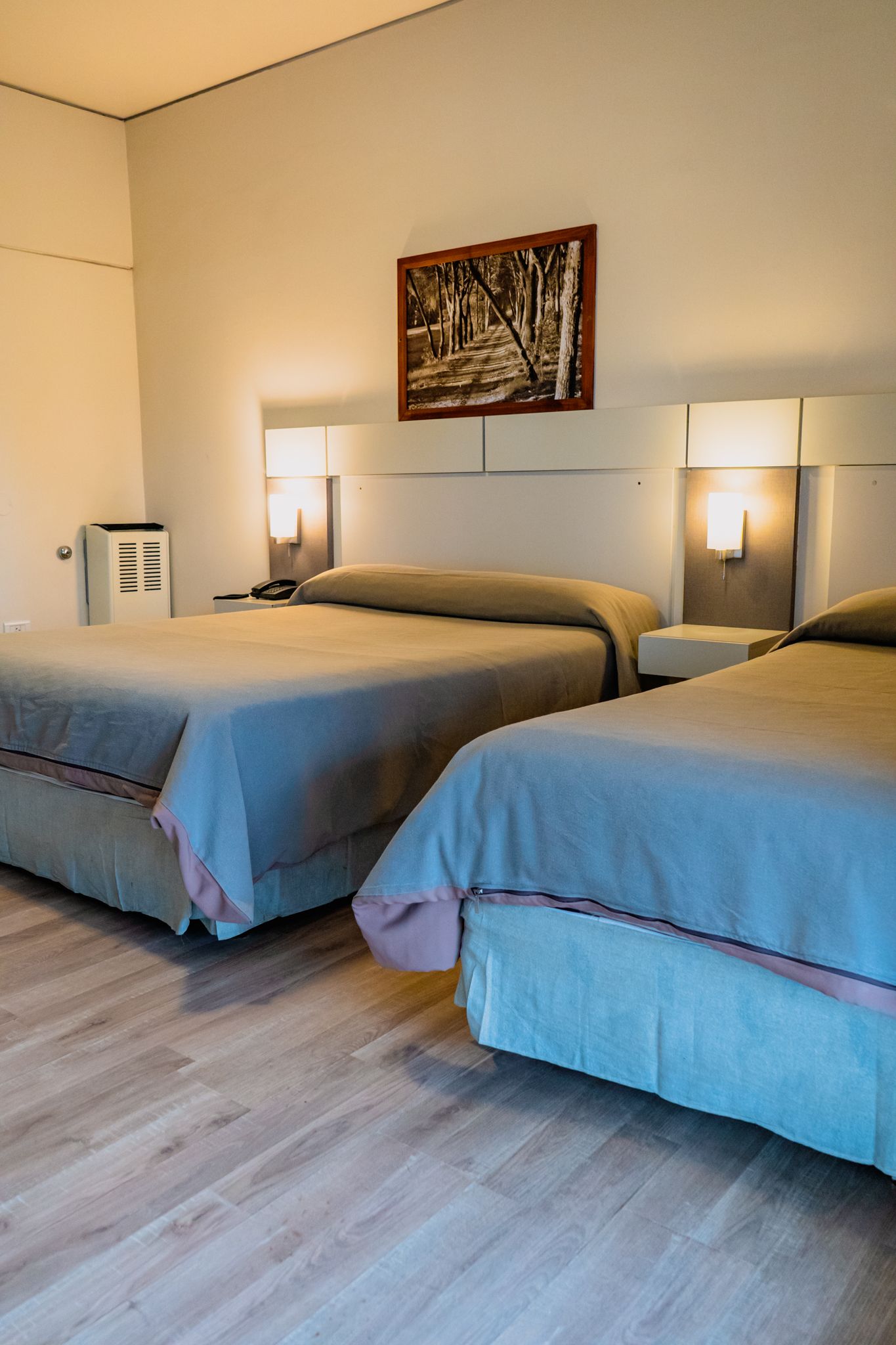 Two beds in a hotel room with gray bedding, wooden floor, neutral walls, and artwork.