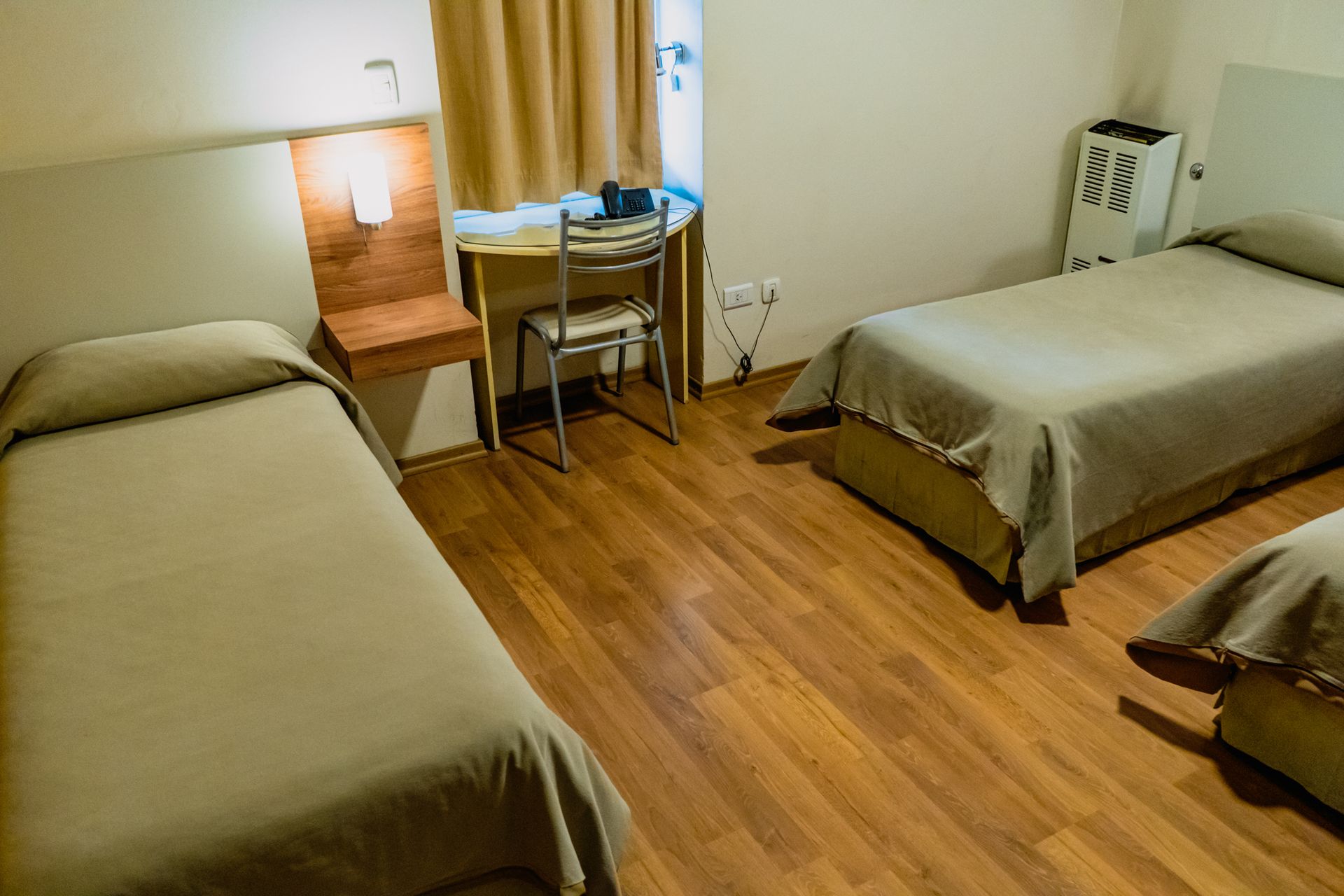 A hotel room with three twin beds, a desk, and a heater. The room has wood flooring.
