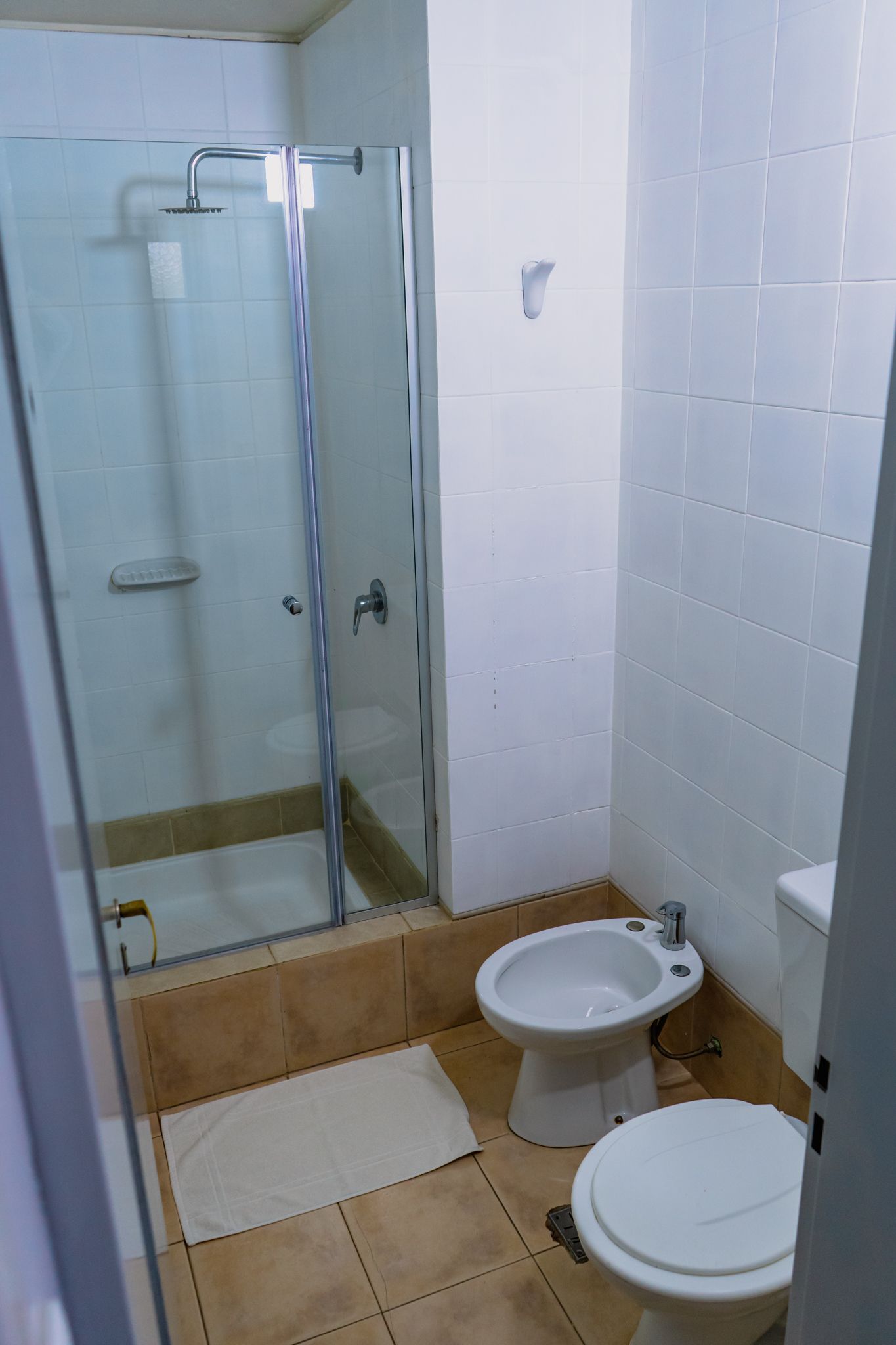 Bathroom with glass shower, toilet, and bidet; beige tiled floor and white walls.