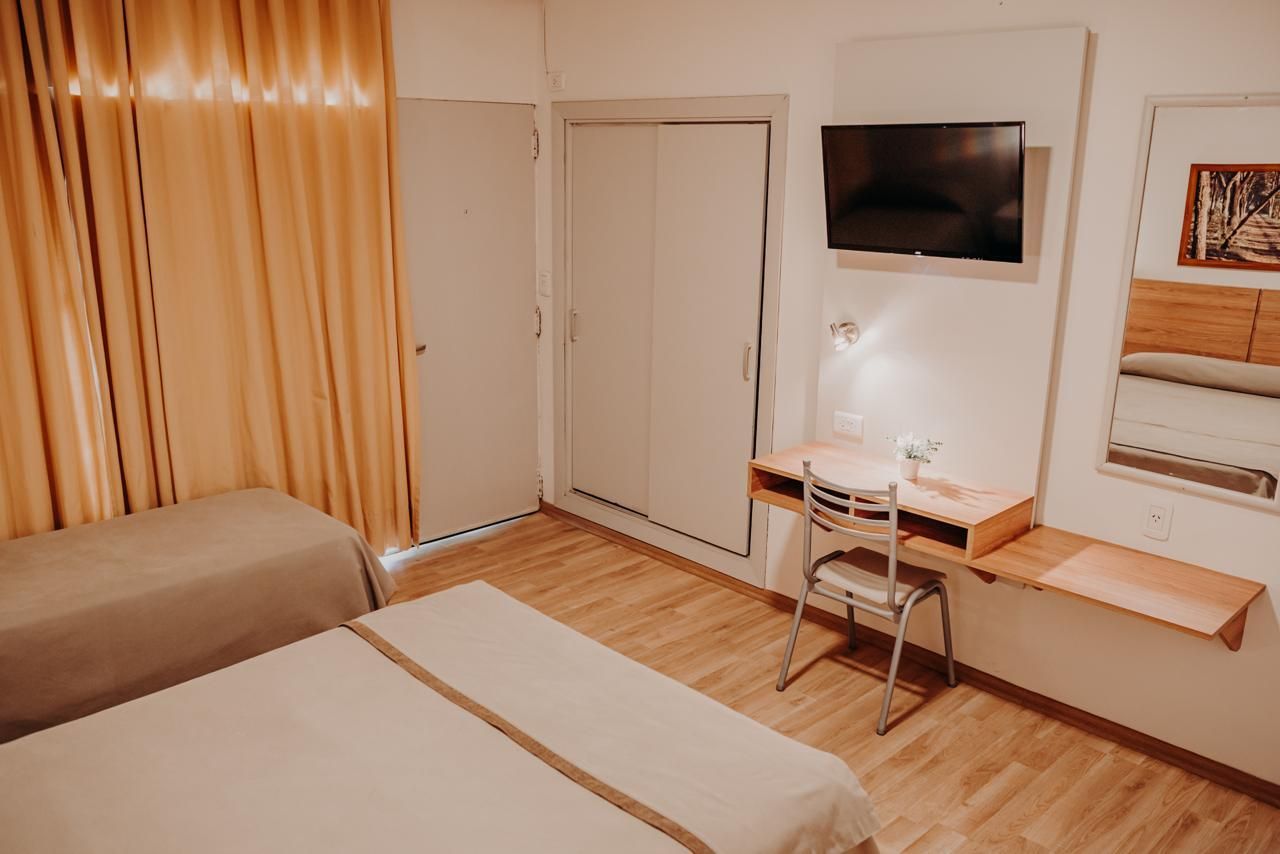 Hotel room with two beds, desk, TV, and closet; beige curtains and wooden floors.