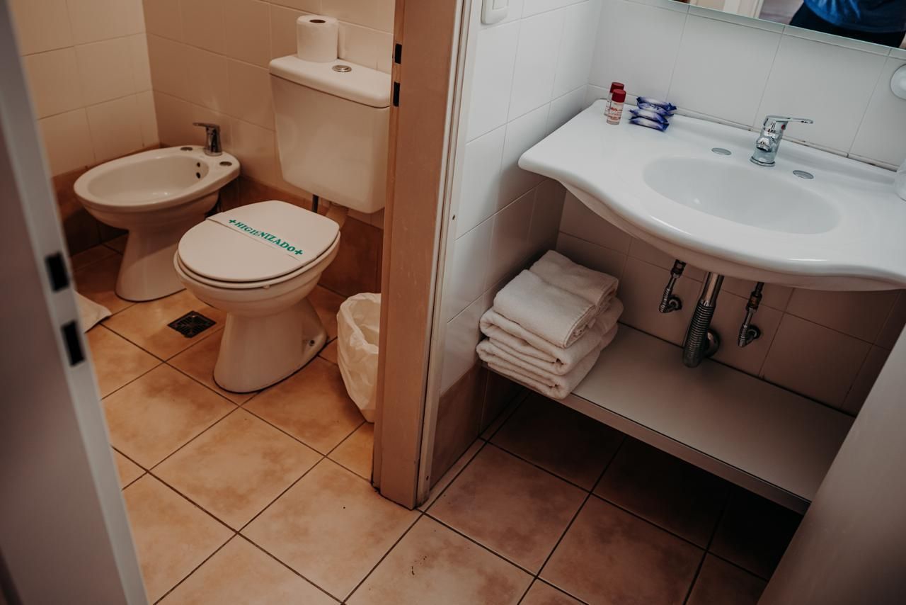 Bathroom with a toilet, bidet, sink, and towels. Tile floor and off-white walls.