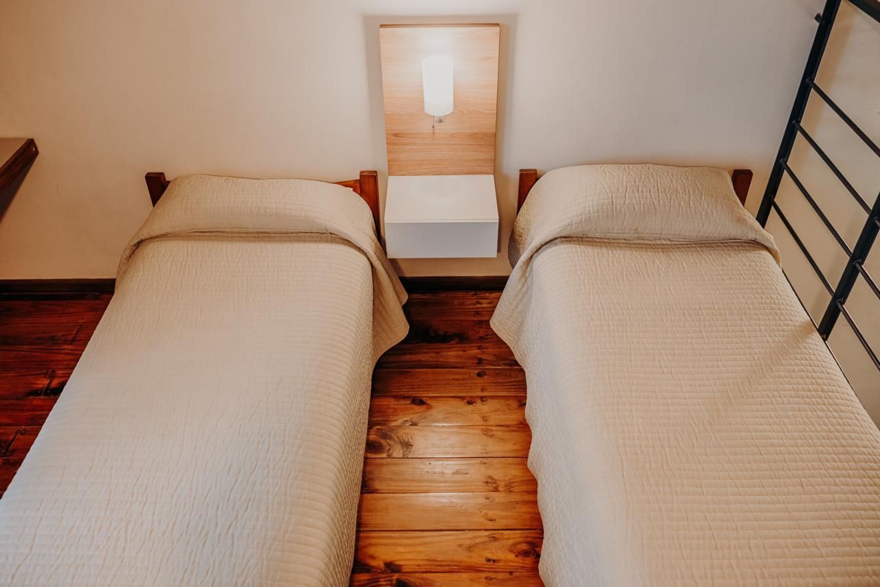 Two beds with beige bedding in a room with wood floors and a wall-mounted light.