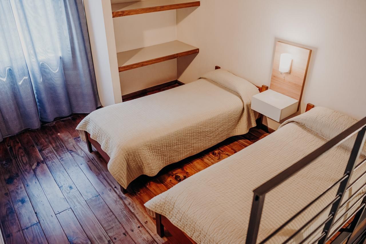 Two twin beds in a small room with wooden floors, shelves, and a window with blue curtains.