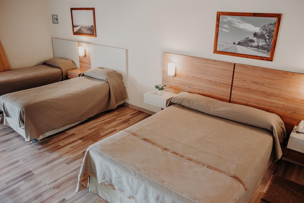 A hotel room with three beds, wooden headboards, neutral bedding, and framed artwork.