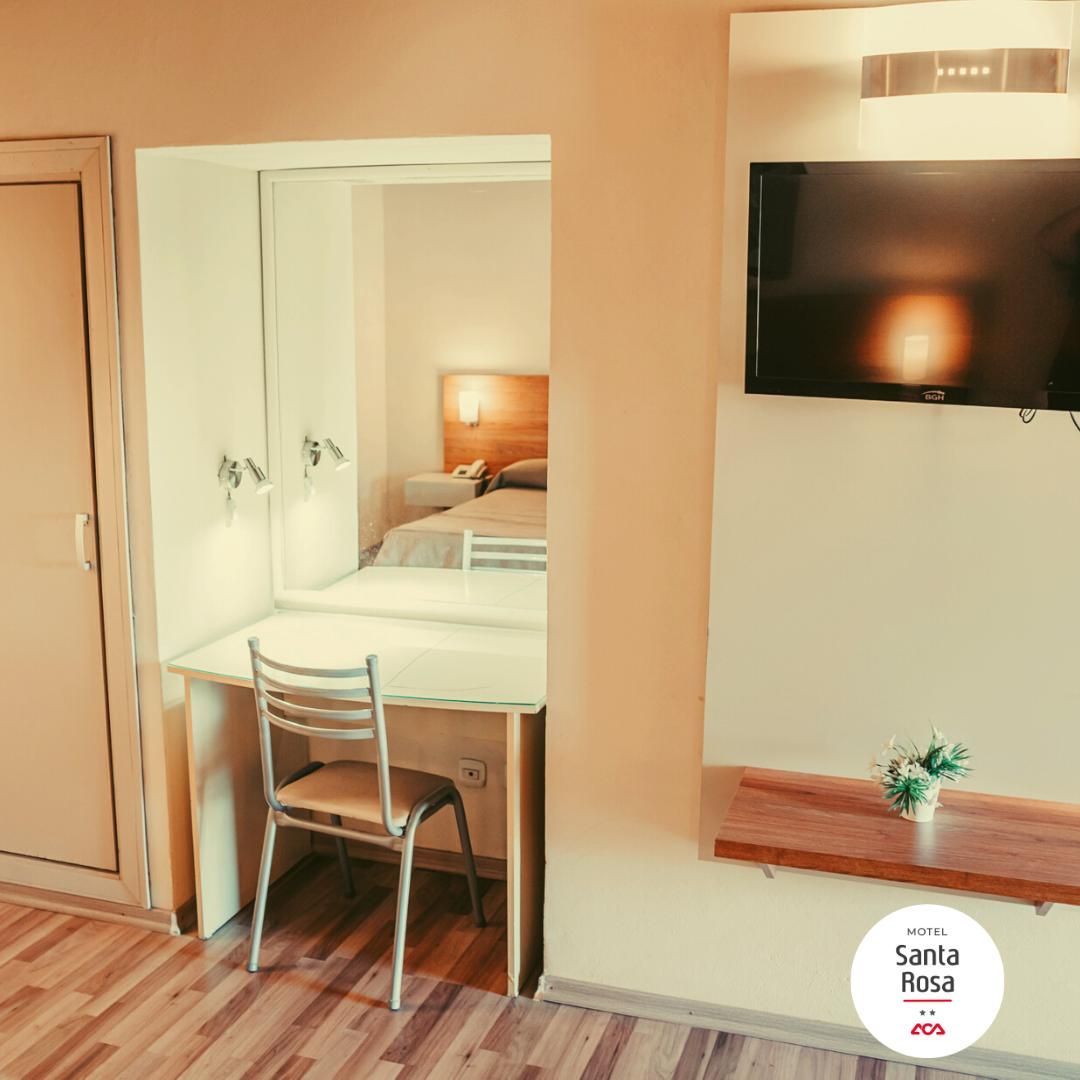 Room with bed, desk, chair, and TV; wood floor, neutral walls, small flowers on shelf.