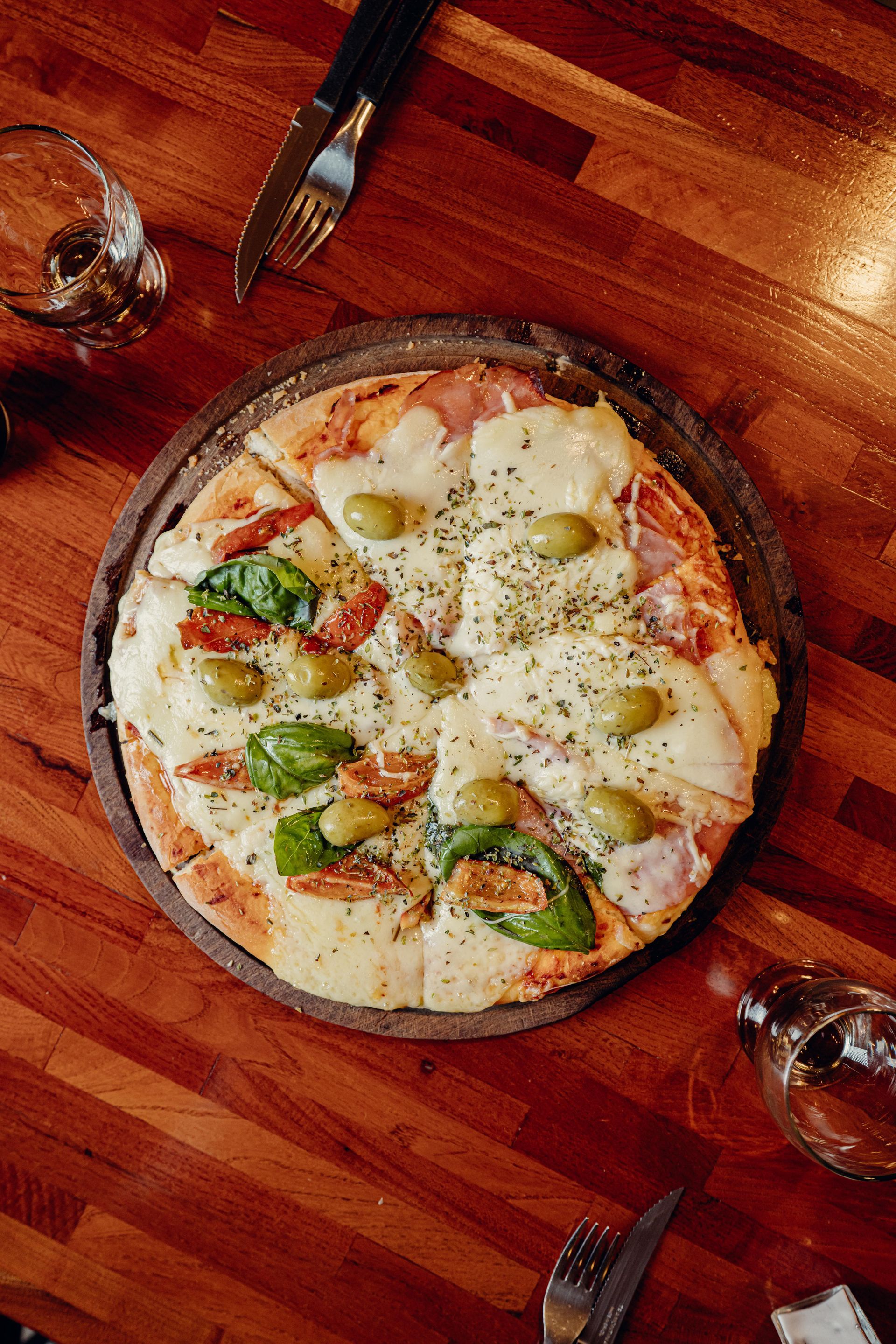Pizza sobre tabla de madera con queso, aceitunas y albahaca sobre una mesa de madera, con cubiertos y vasos.