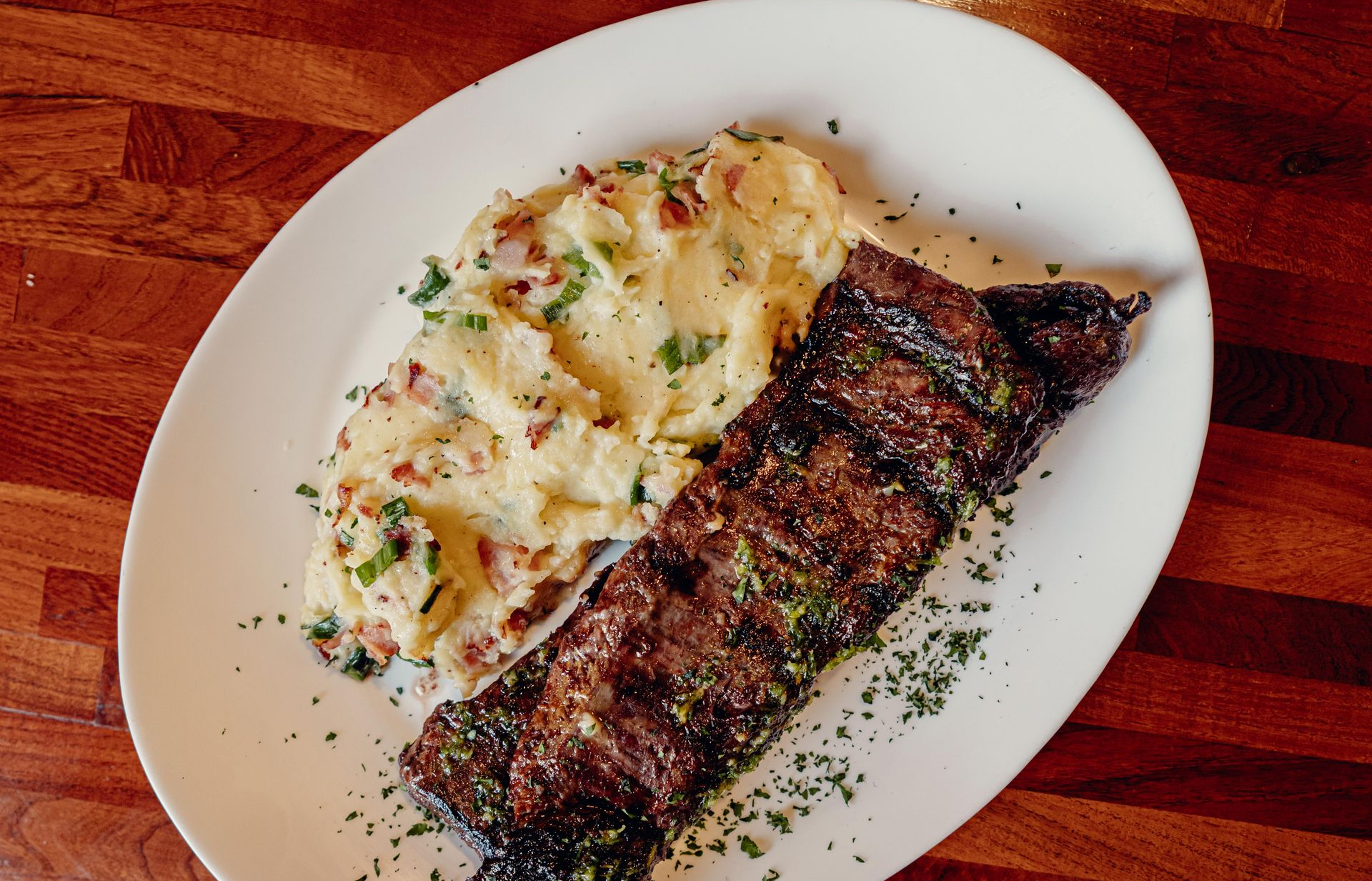 Bistec a la parrilla y puré de papas en un plato blanco, con guarnición de perejil, servido sobre madera.
