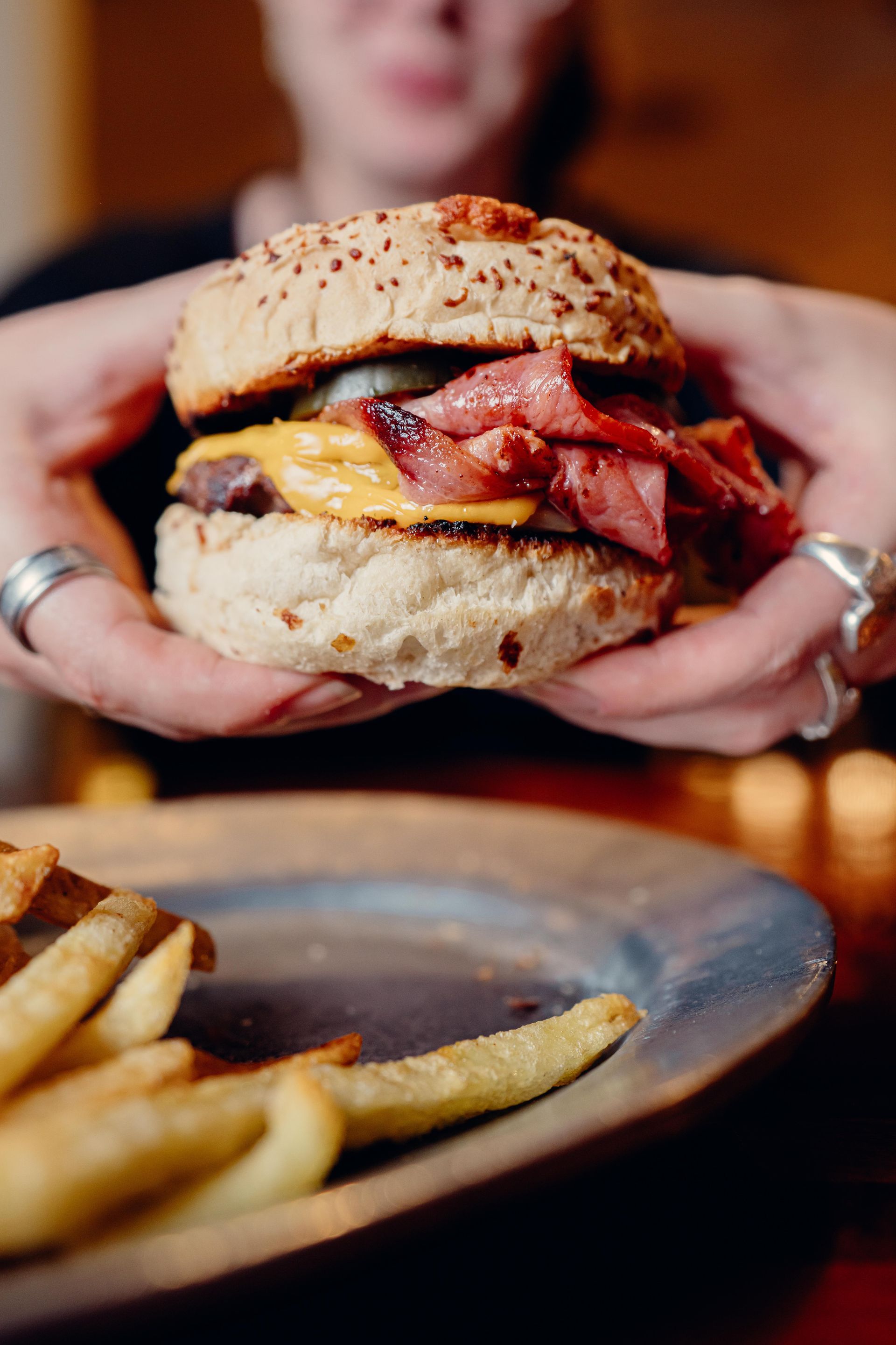 Persona sosteniendo una hamburguesa grande con tocino, queso y un pan de sésamo, junto a papas fritas en un plato.