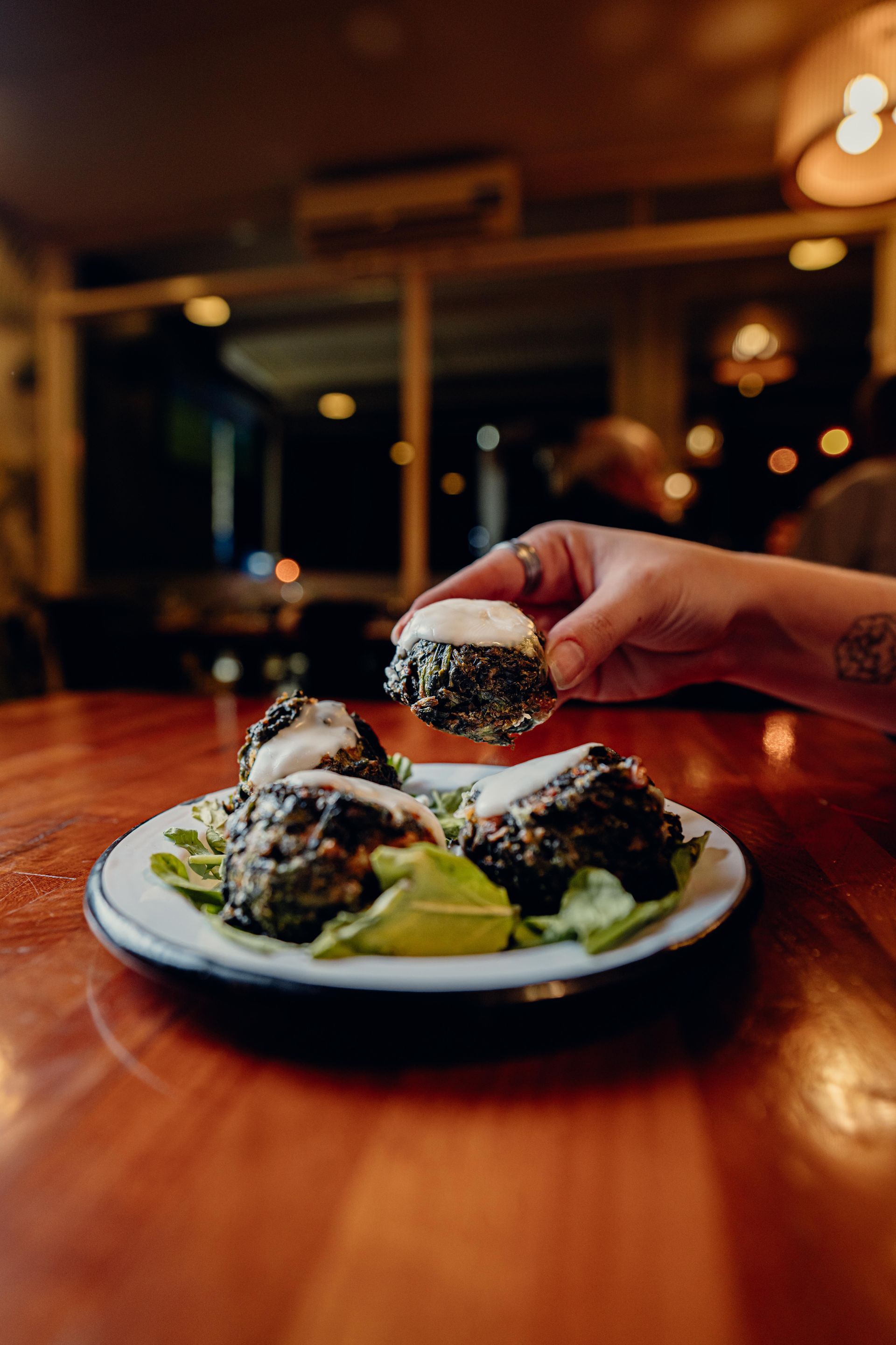 Mano sosteniendo un alimento cubierto de semillas de sésamo sobre un plato con elementos similares en una mesa de madera.