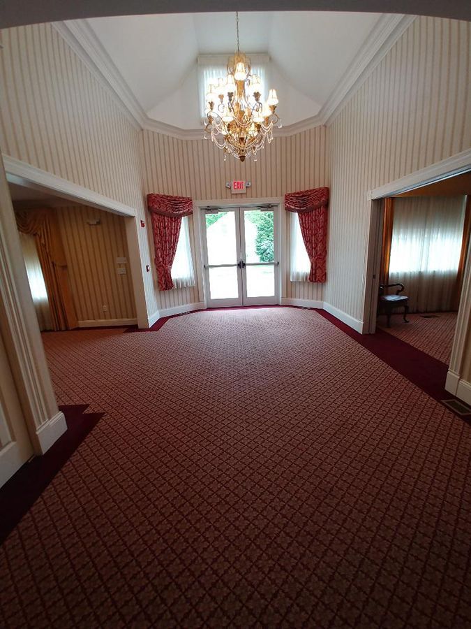 Lobby with red patterned carpet, chandelier, and double doors. Red curtains frame the doors. Light-colored walls.