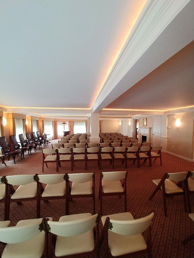 Rows of chairs in a room with a cross, likely a funeral home chapel, with neutral walls and red carpet.