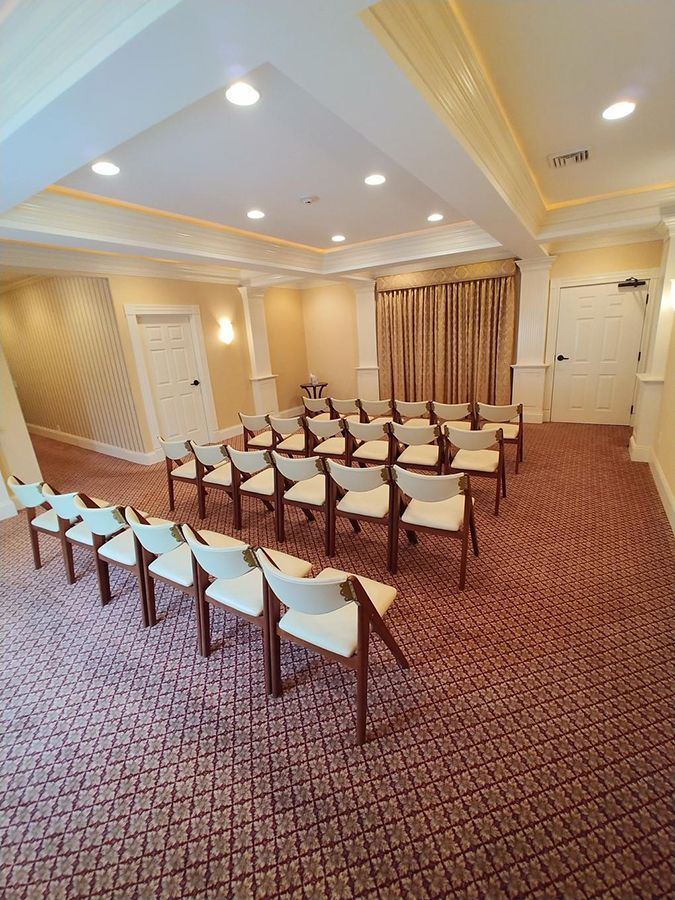 Rows of chairs in a room, likely for a meeting or event. Beige walls, red carpet, neutral chairs, curtains.