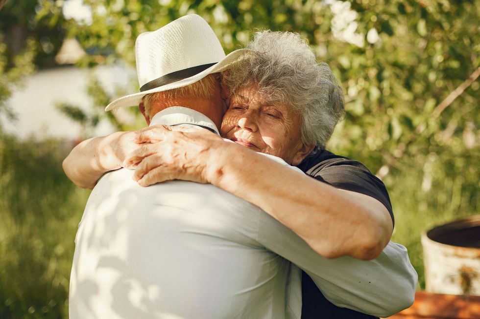 Two people hugging outdoors in a sunny garden, one wearing a white hat.