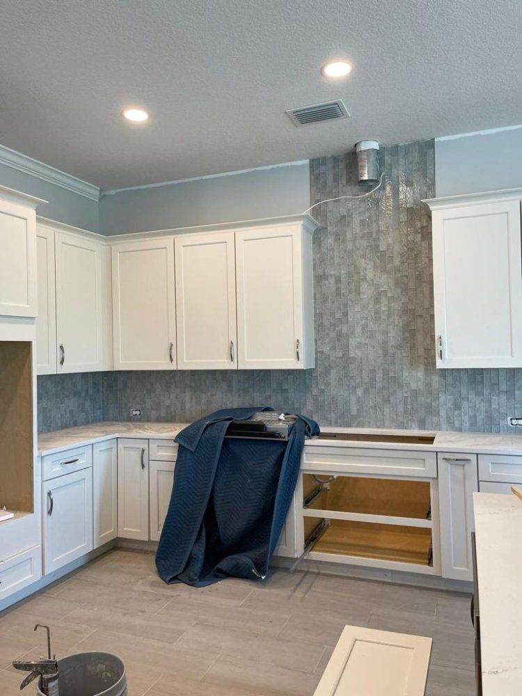 White kitchen cabinets, blue-gray tile backsplash, light blue walls, and light gray flooring in a room under construction.