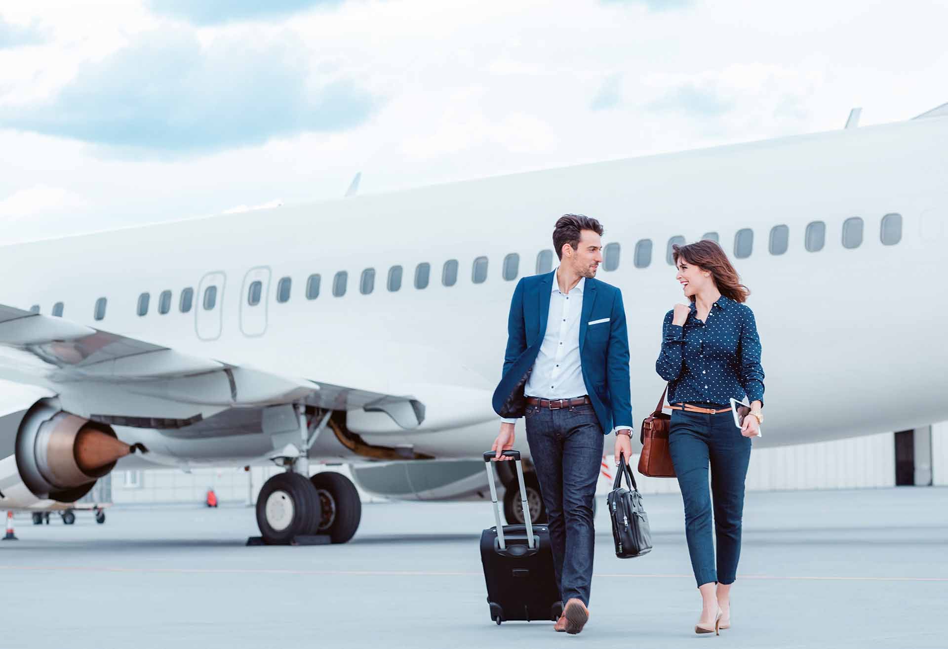 Couple walking towards a plane on the tarmac, man rolling a suitcase, woman carrying a purse, bright sky.