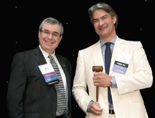 Two men, one holding a gavel, in suits with name tags, smiling in front of a black backdrop.