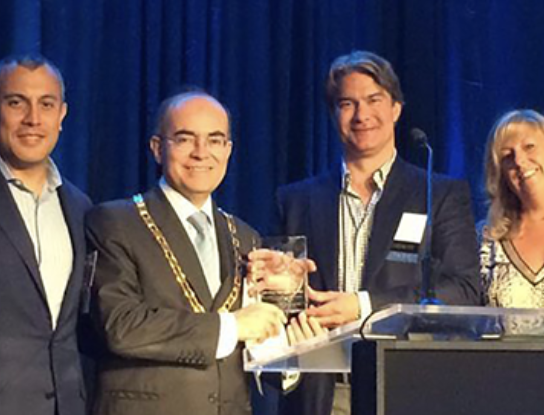Men and woman on stage, one man holding an award. Blue background, podium.
