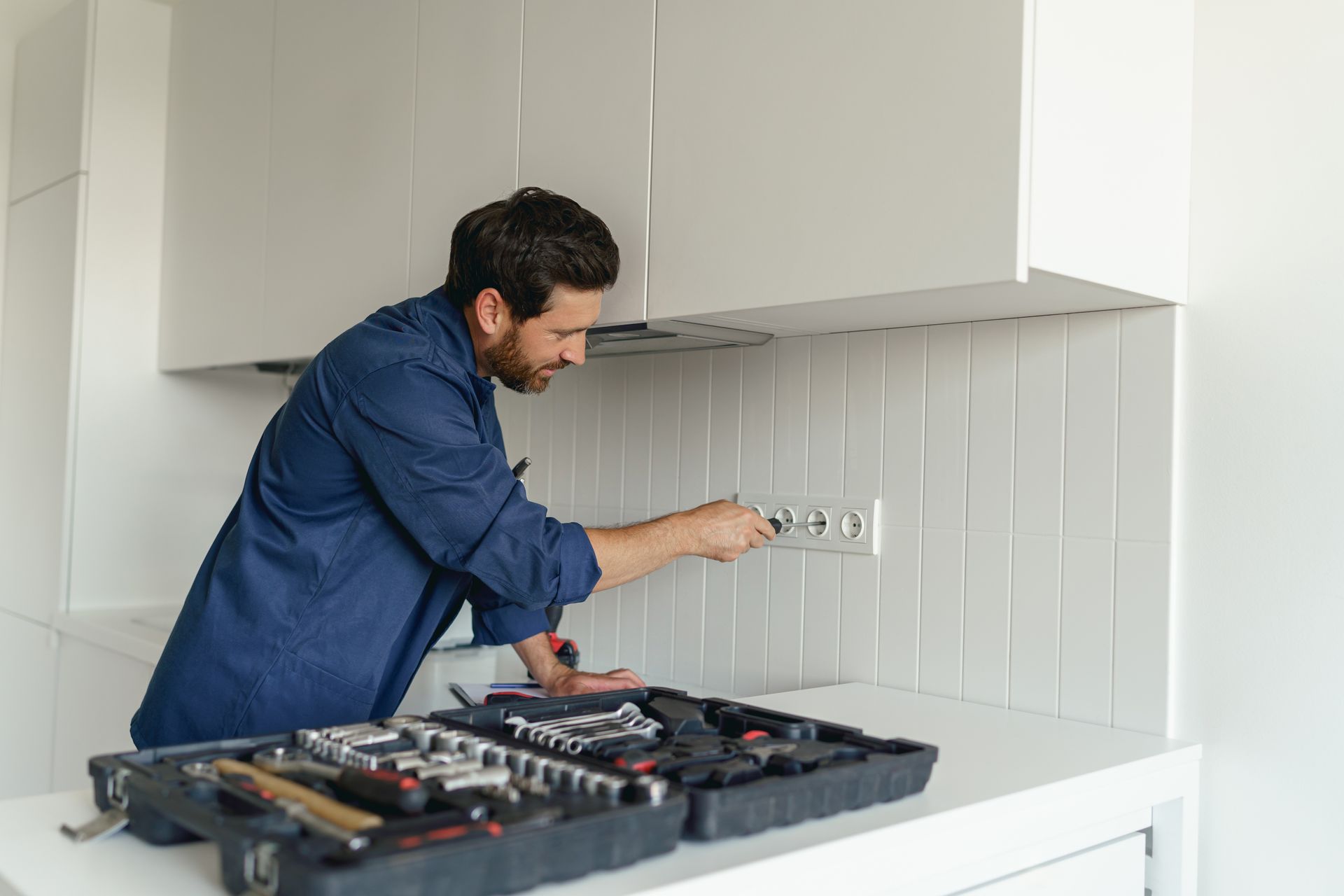A person in a blue shirt works on an electrical wall outlet in a white kitchen, with a tool kit open on the counter.