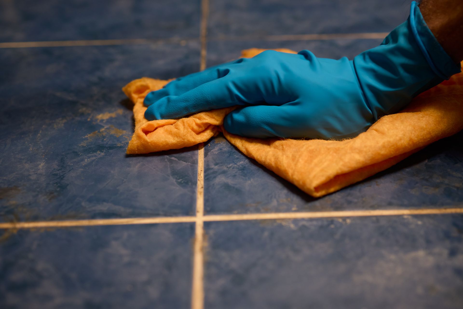 A gloved hand uses an orange cloth to wipe down blue tiled flooring.