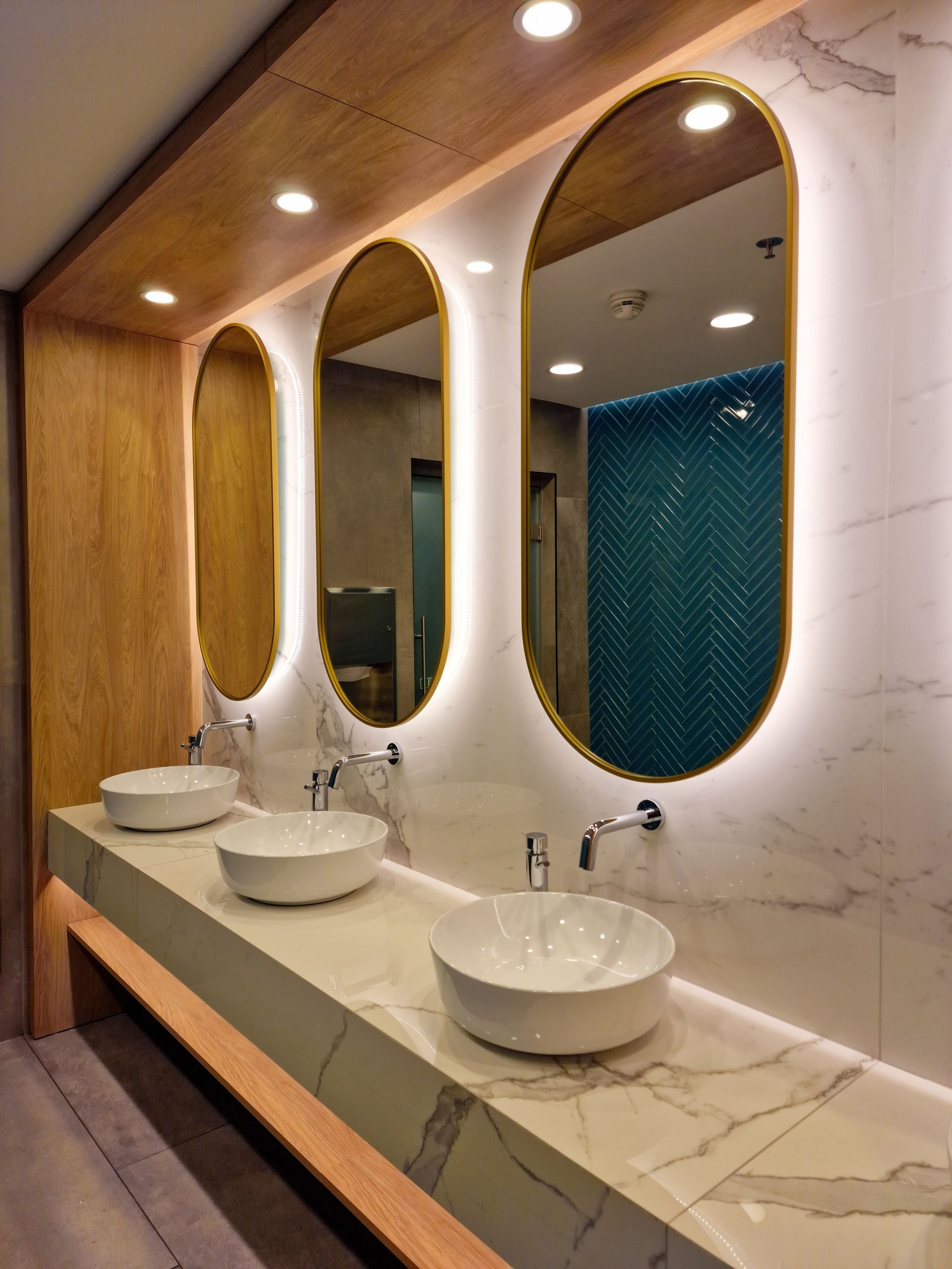 Modern bathroom vanity with three oval backlit mirrors, white vessel sinks, and a marble countertop against a blue wall.