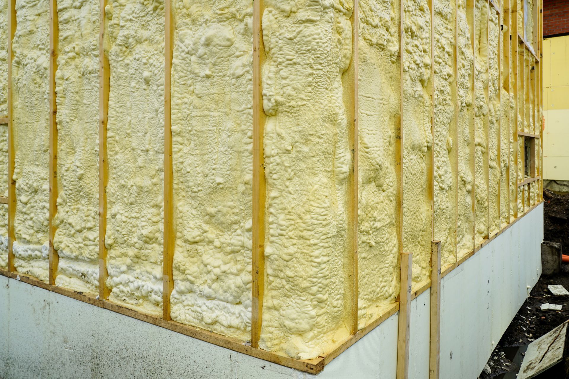 Spray foam insulation applied between wooden wall studs above a white foundation base on a construction site.