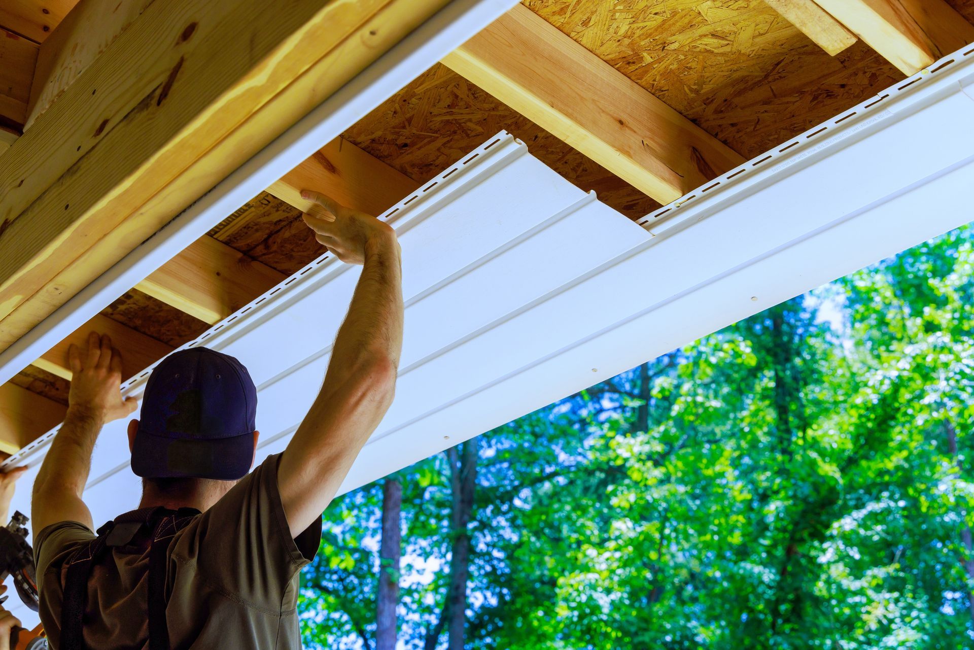 A person in a dark cap installs white vinyl soffit panels to the underside of a wooden roof overhang.