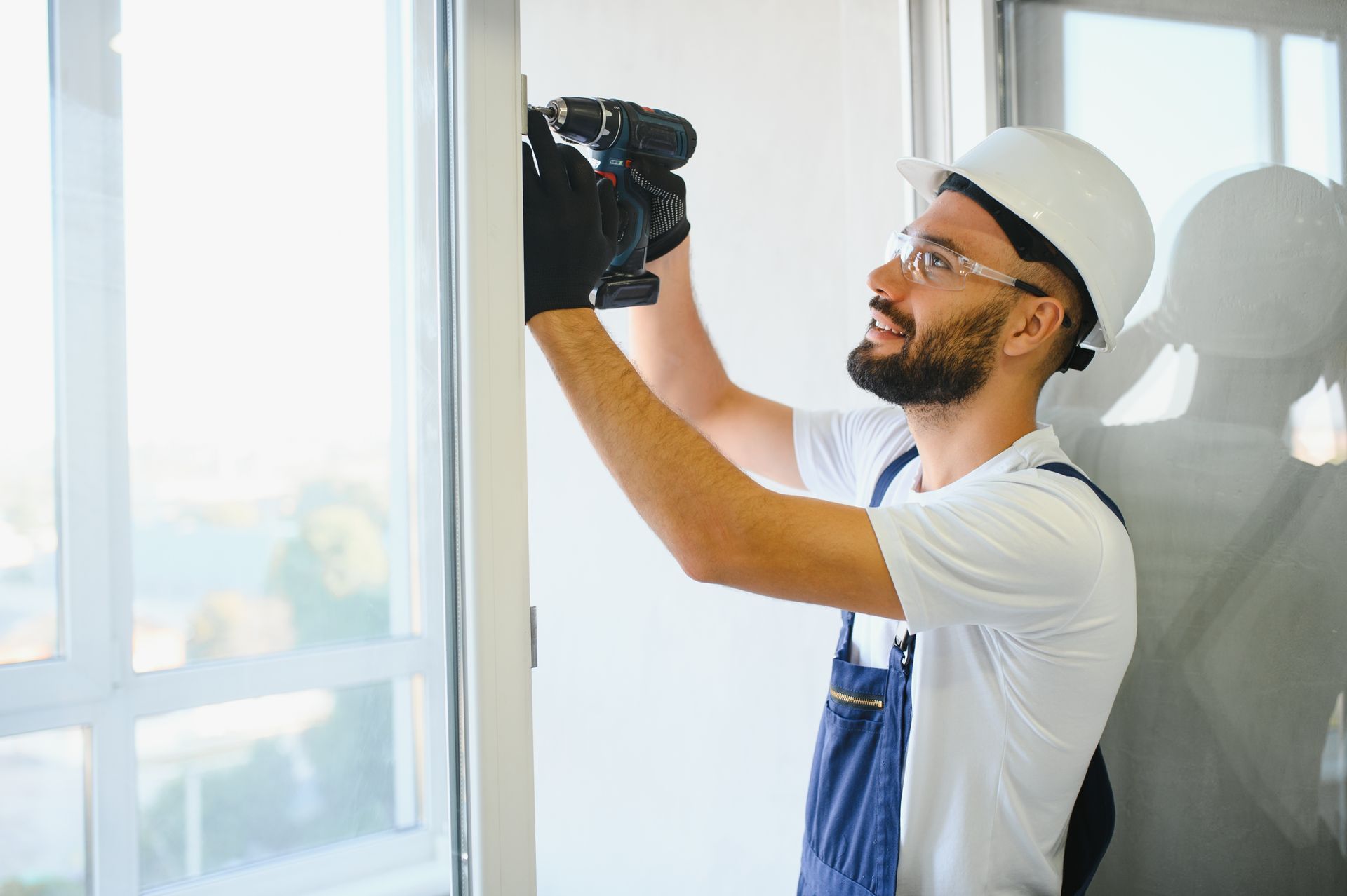A worker in a white hard hat and blue coveralls uses a power drill to install a frame near a large window.